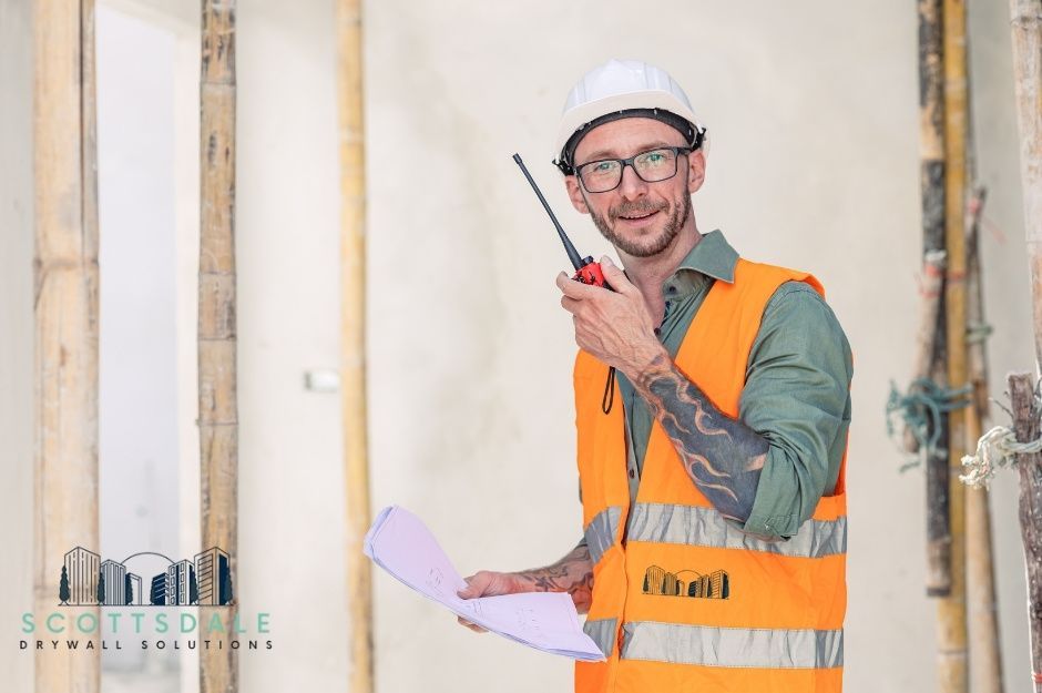 A drywall repair handyman wearing a white hard hat, glasses, and an orange high-visibility vest over a green shirt holding a walkie-talkie radio and blueprints. In a commercial site with wooden framing near W Lawrence Ln, and N Lizanne Way, Tolleson, AZ.