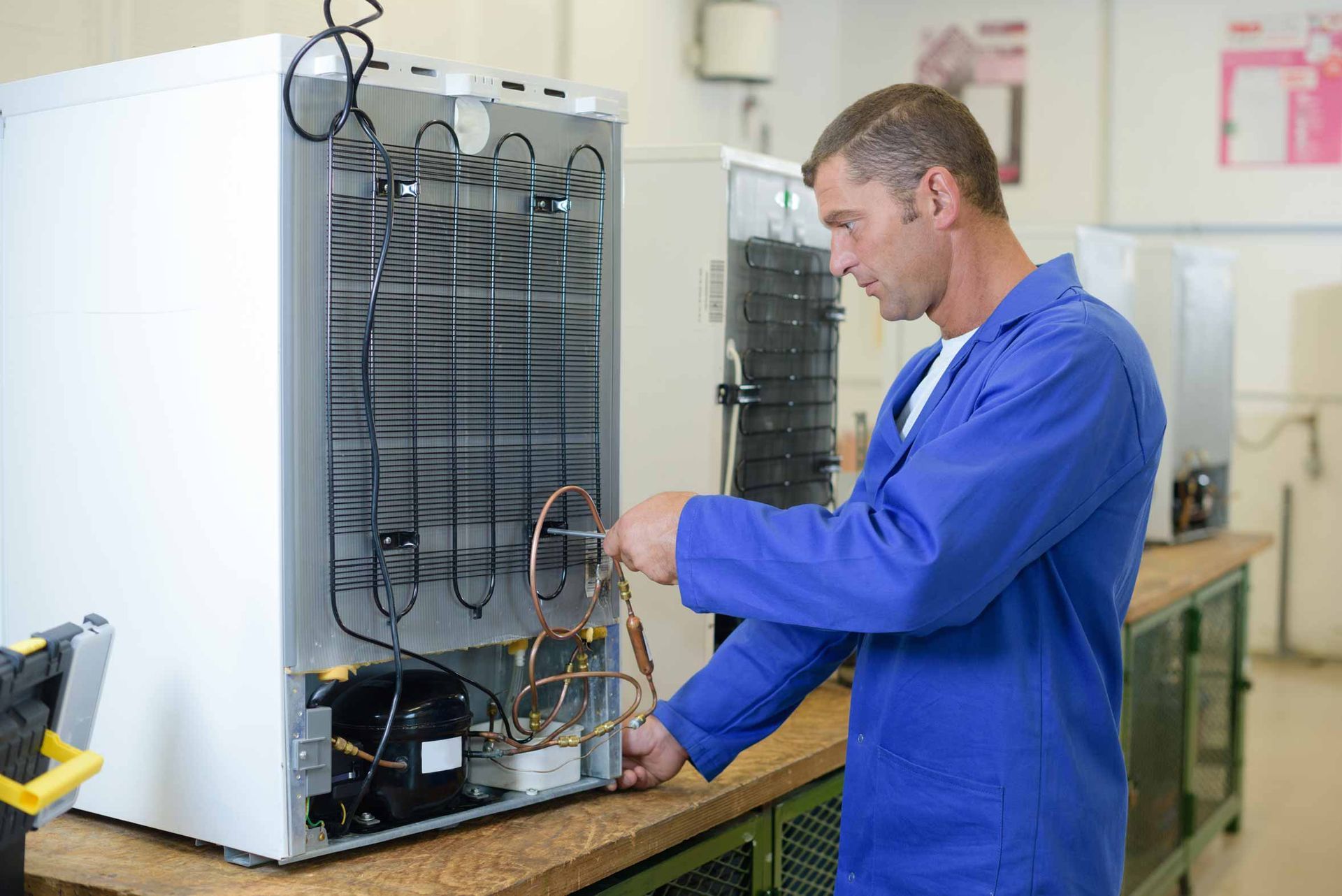 Technician in blue coveralls inspecting the back of a refrigerator on a workbench