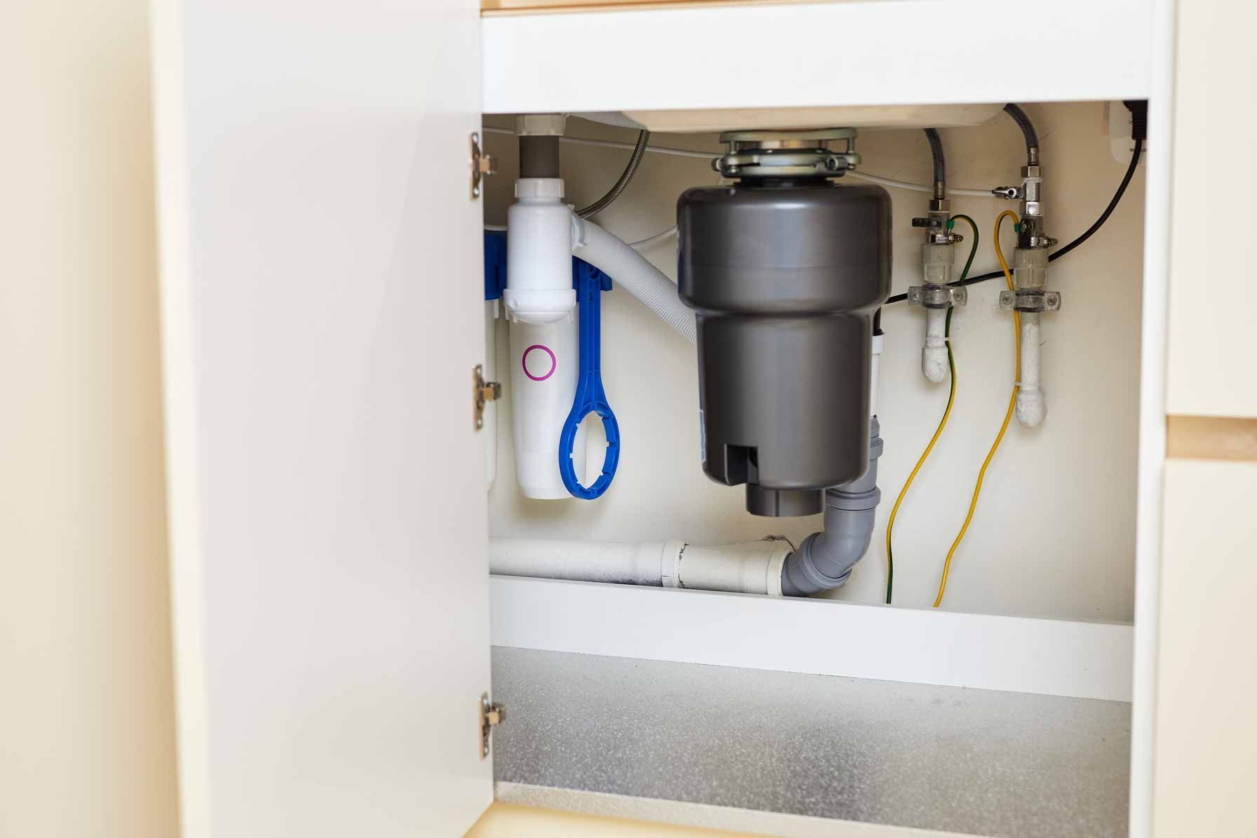 Open kitchen cabinet with plumbing pipes, valves, and a gray garbage disposal unit.