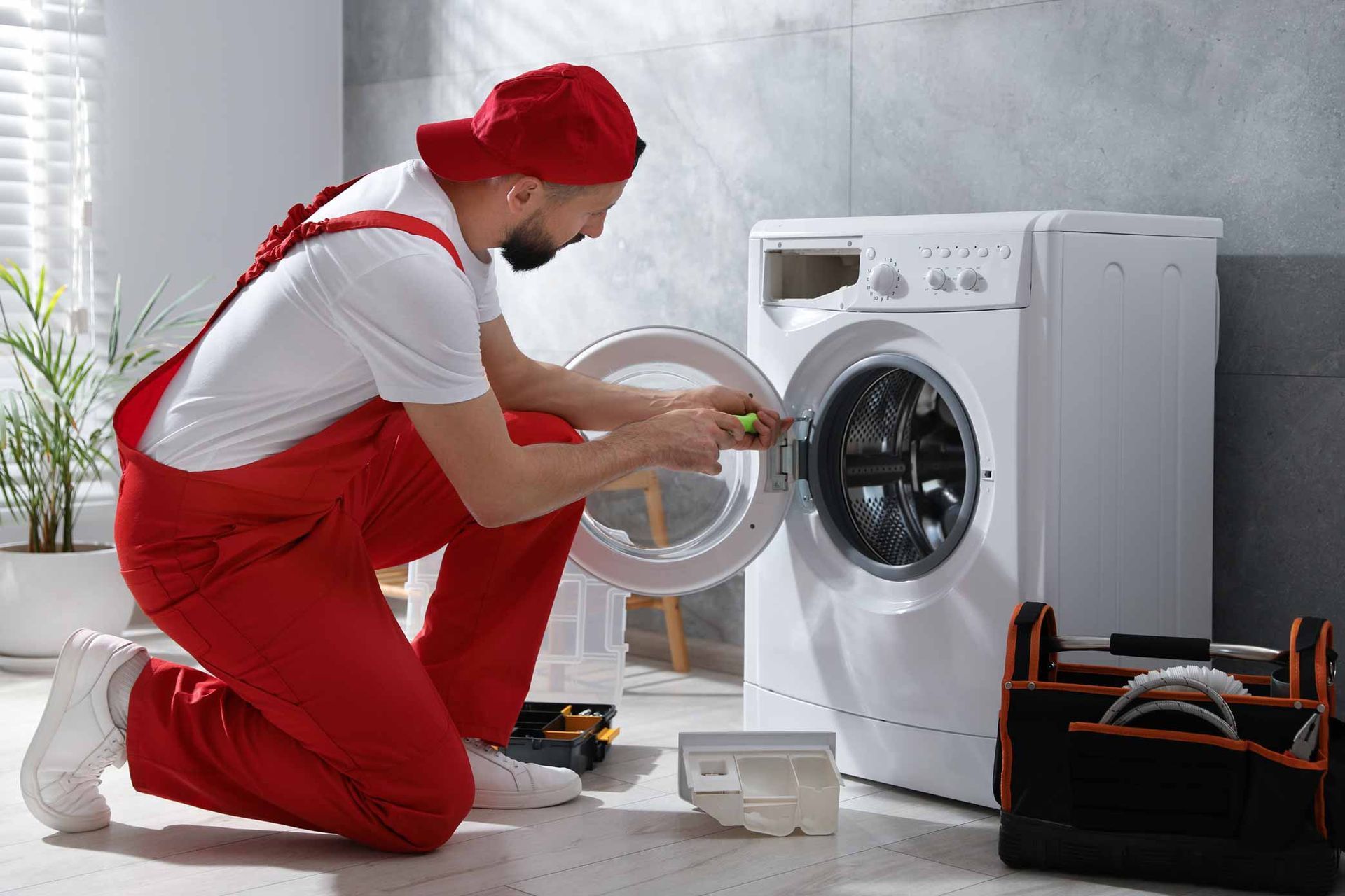 Technician in red overalls repairing a front-loading washing machine in a bright laundry room