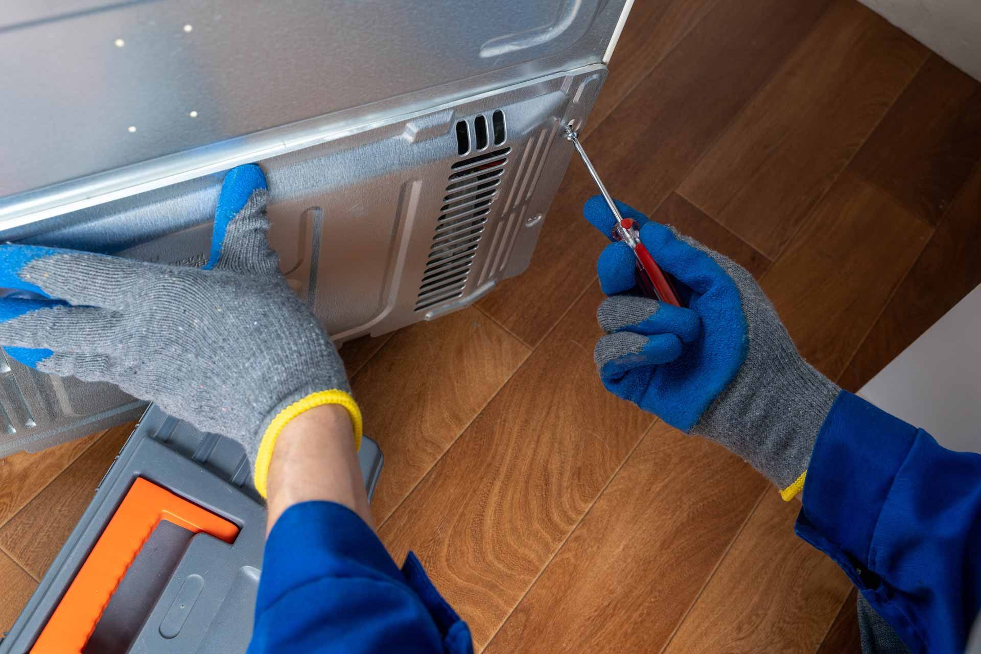 Hands in gray and blue gloves inspecting a silver appliance with a screwdriver on a wooden floor