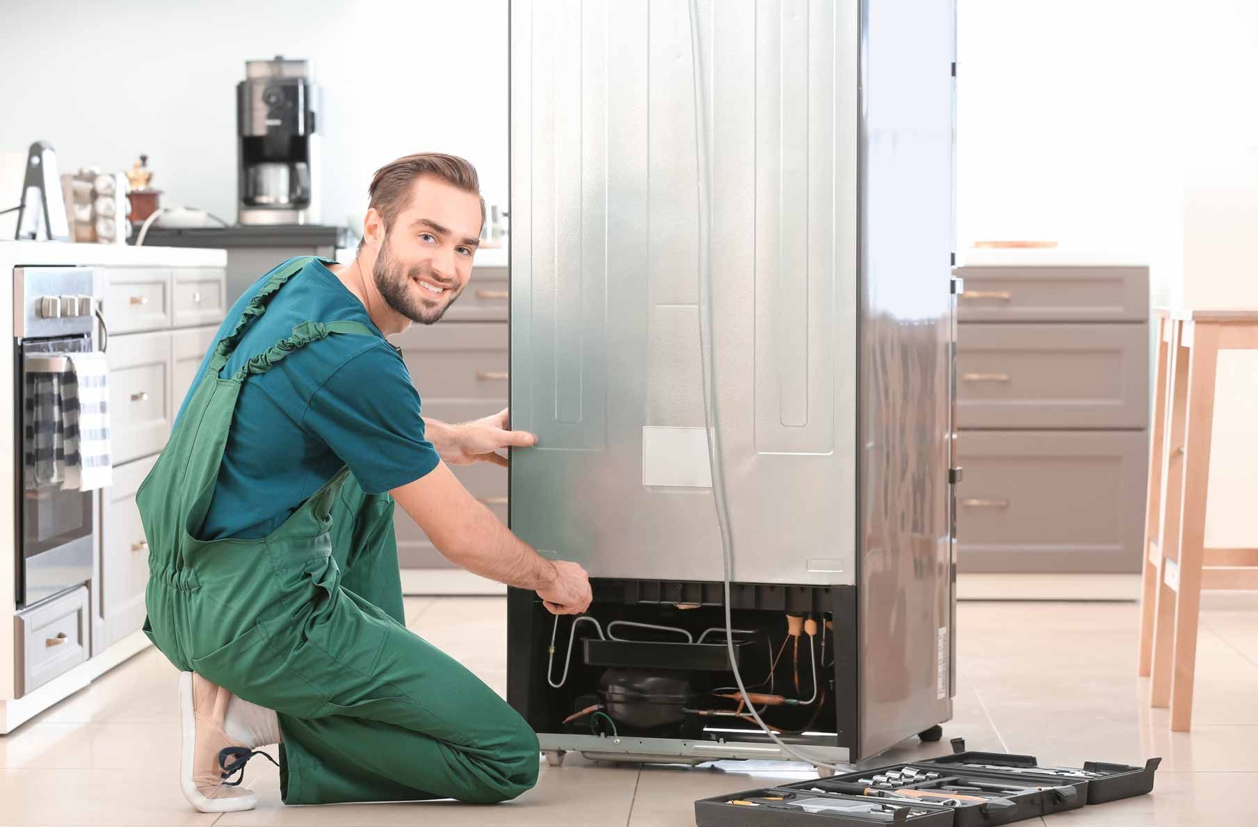 Man in green overalls kneeling to repair a refrigerator in a bright kitchen.