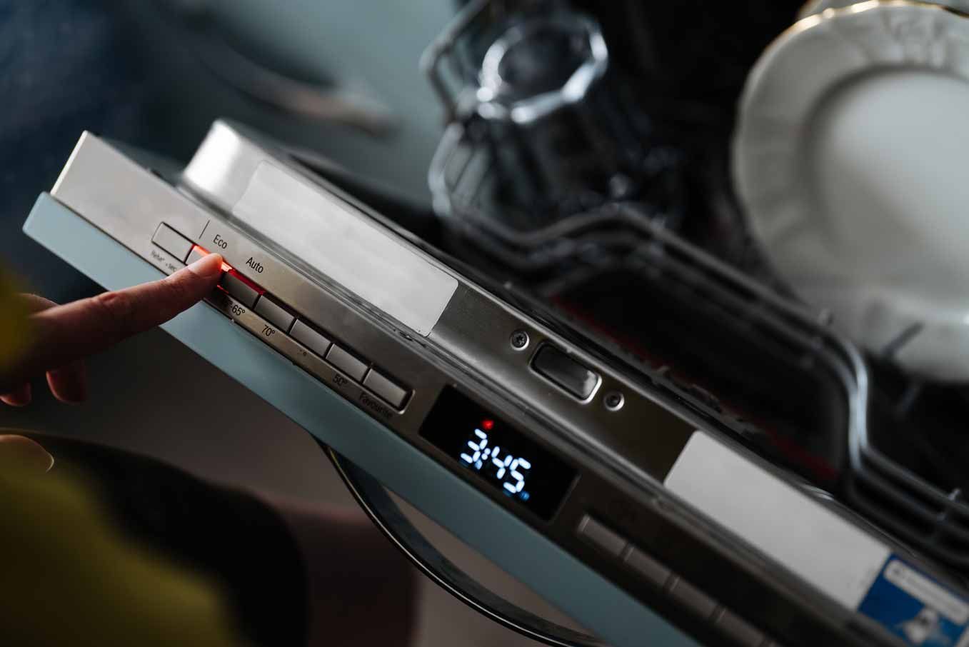 Hand pressing a dishwasher control panel with a small blue digital display showing 3:45