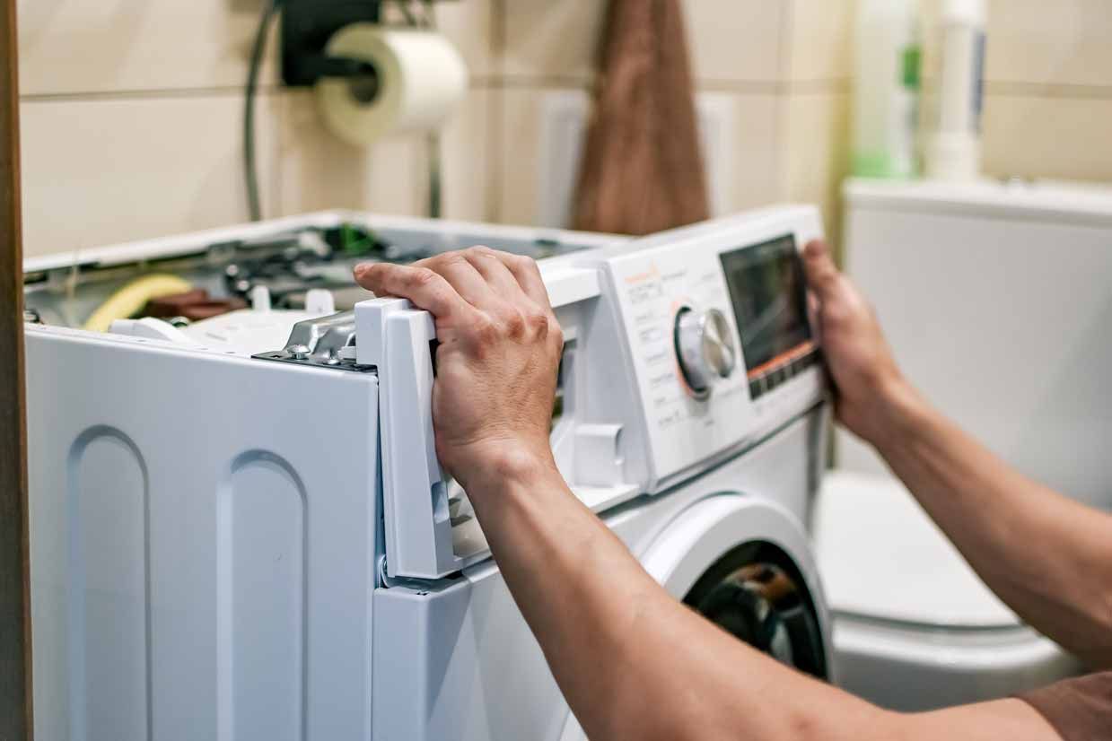 Hands lifting a washing machine lid in a laundry room, with a repair panel open.