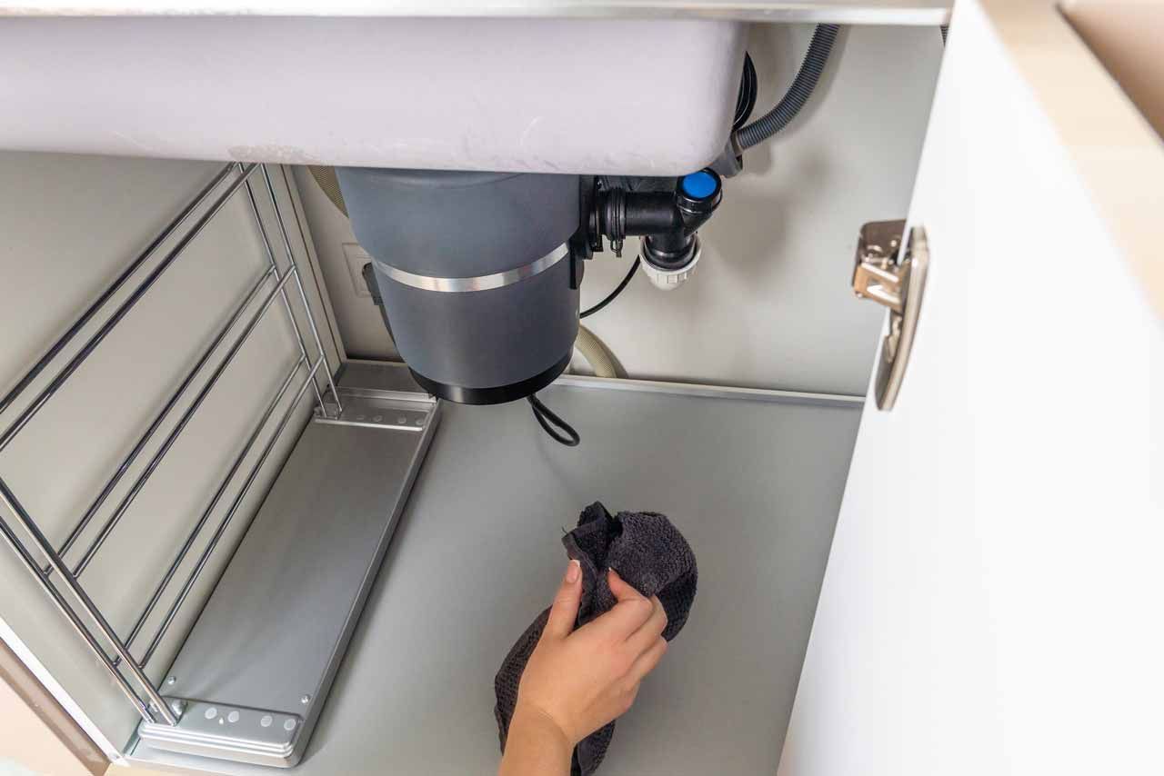 Hand reaching into a cabinet under a sink, cleaning a gray basin with a dark cloth.