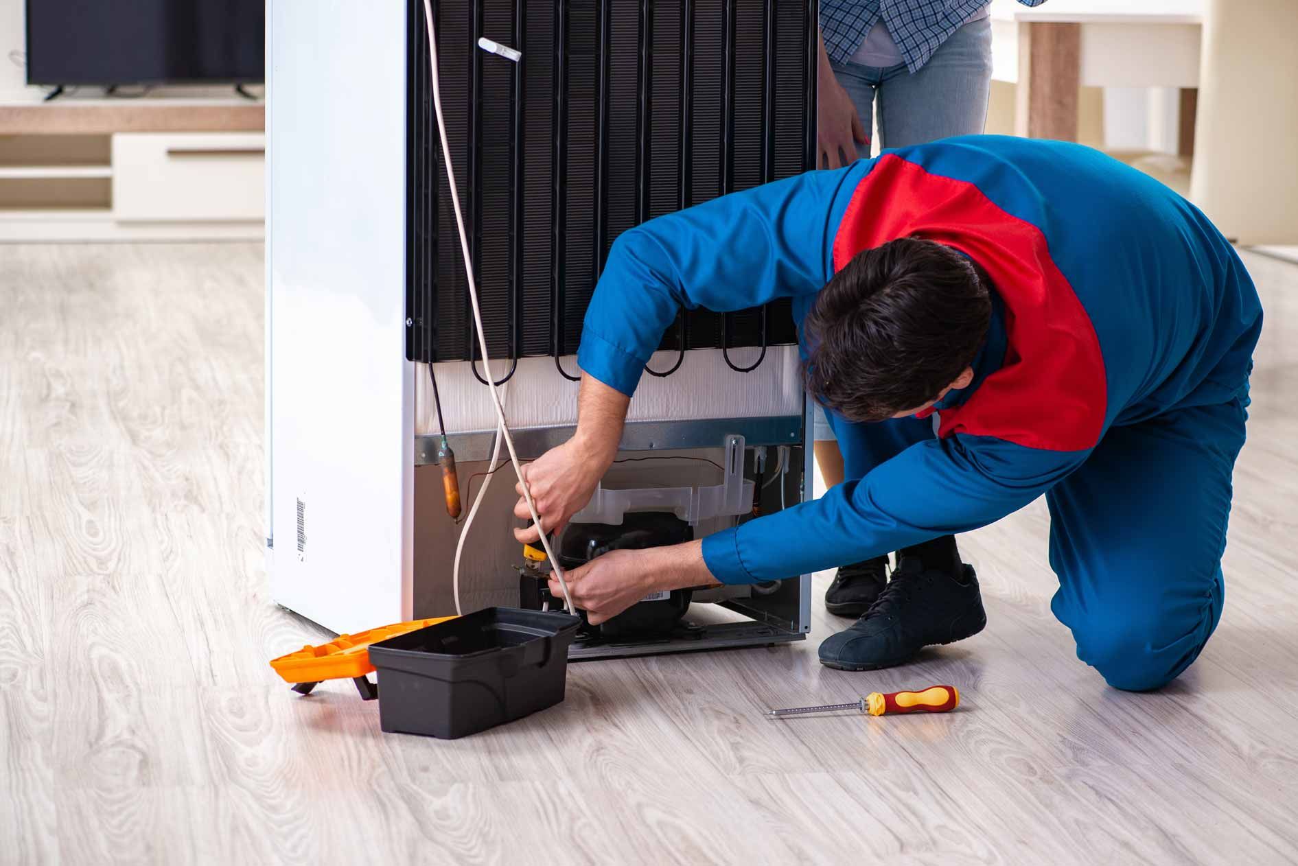 Technician kneeling by a refrigerator, using tools to repair it on a kitchen floor.