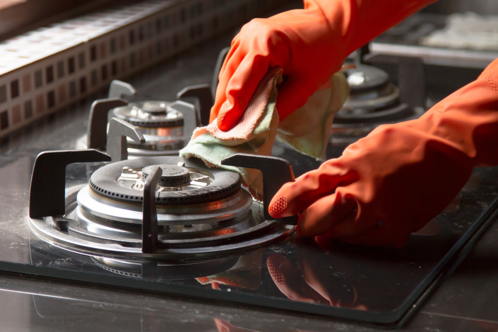 Hands in orange gloves cleaning a gas stovetop with a cloth