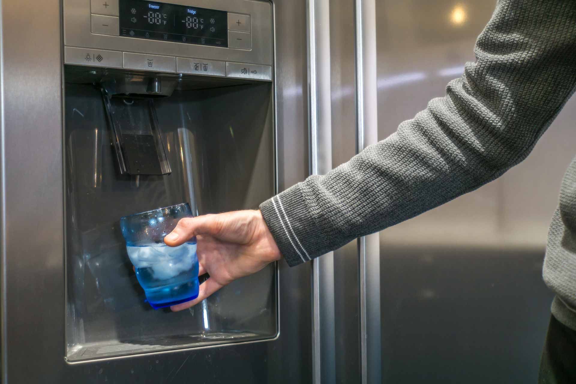 Hand fills a blue-tinted glass with water from a stainless steel refrigerator dispenser