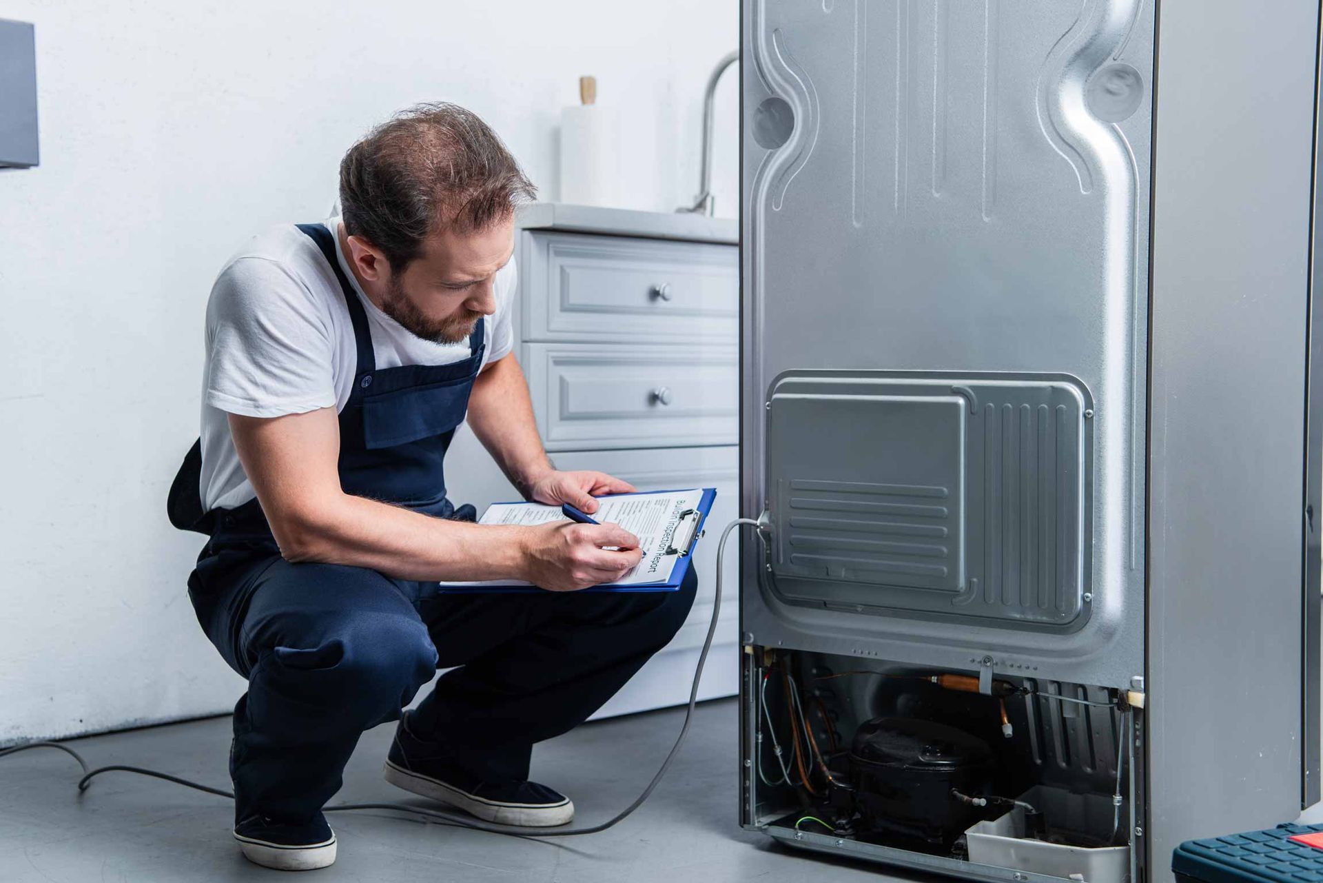 Technician crouches beside a broken appliance, inspecting it with a clipboard in a bright utility room