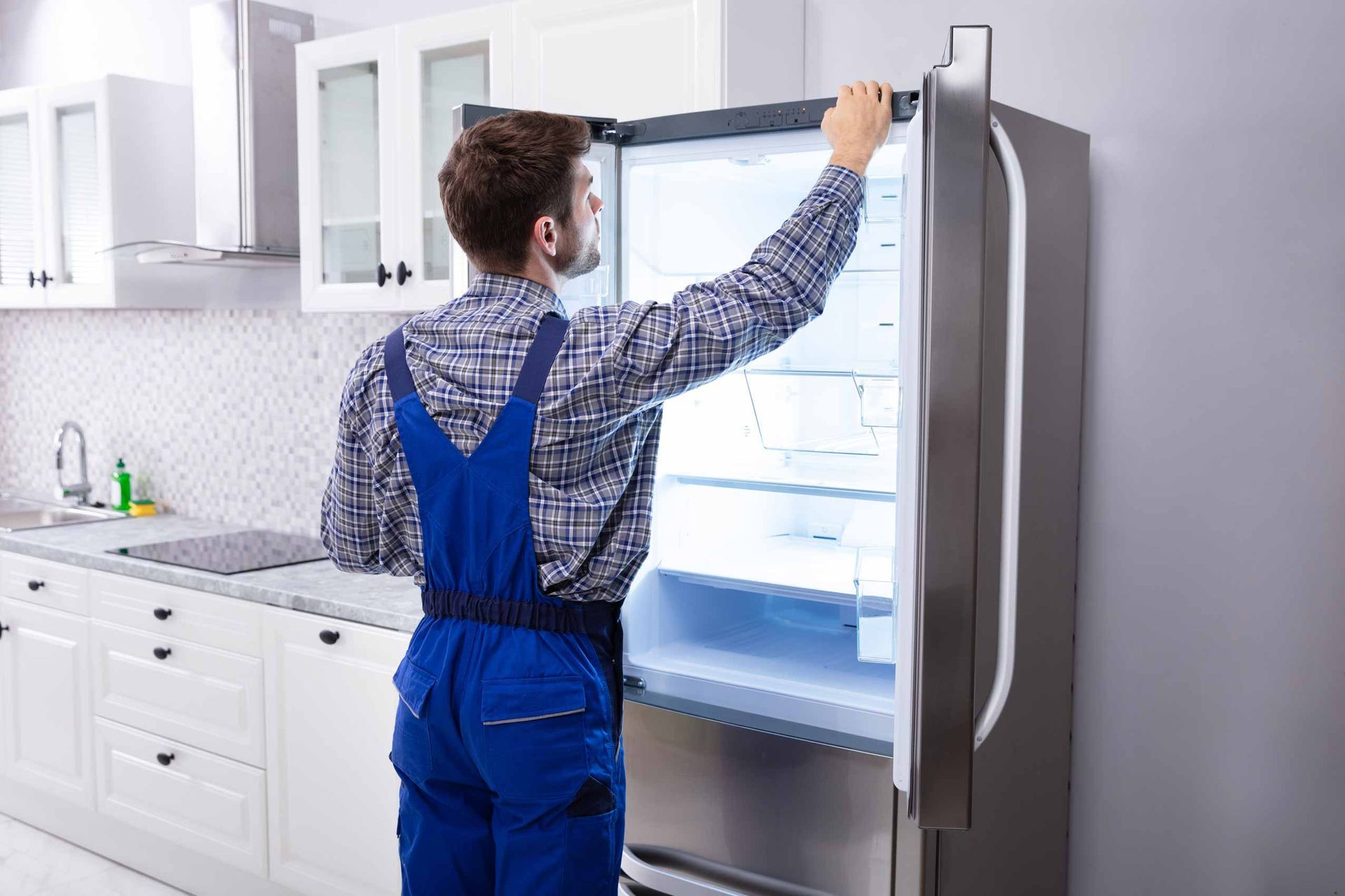 Person in blue overalls reaching into an open refrigerator in a bright kitchen