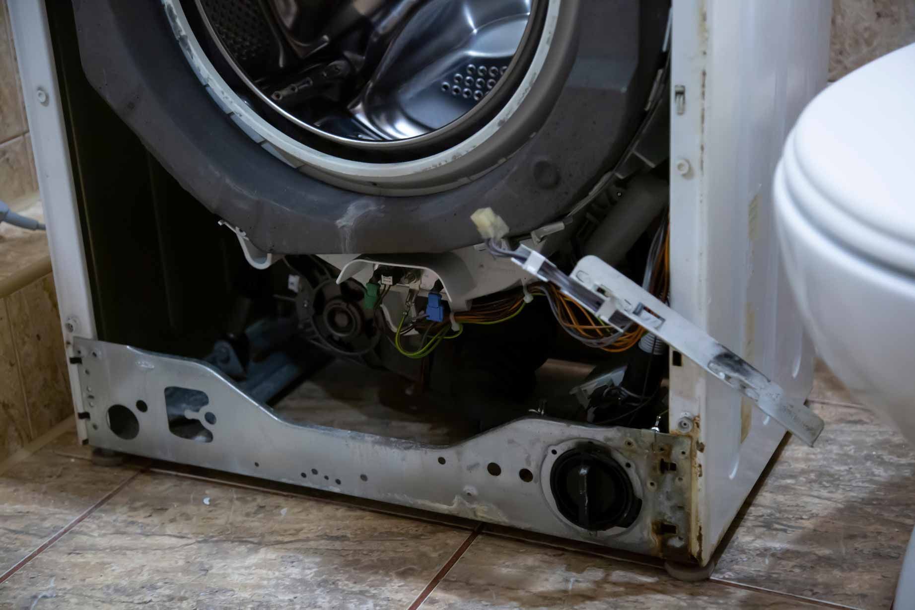 Broken front panel of a white washing machine, with exposed inner drum and wiring on a tiled floor