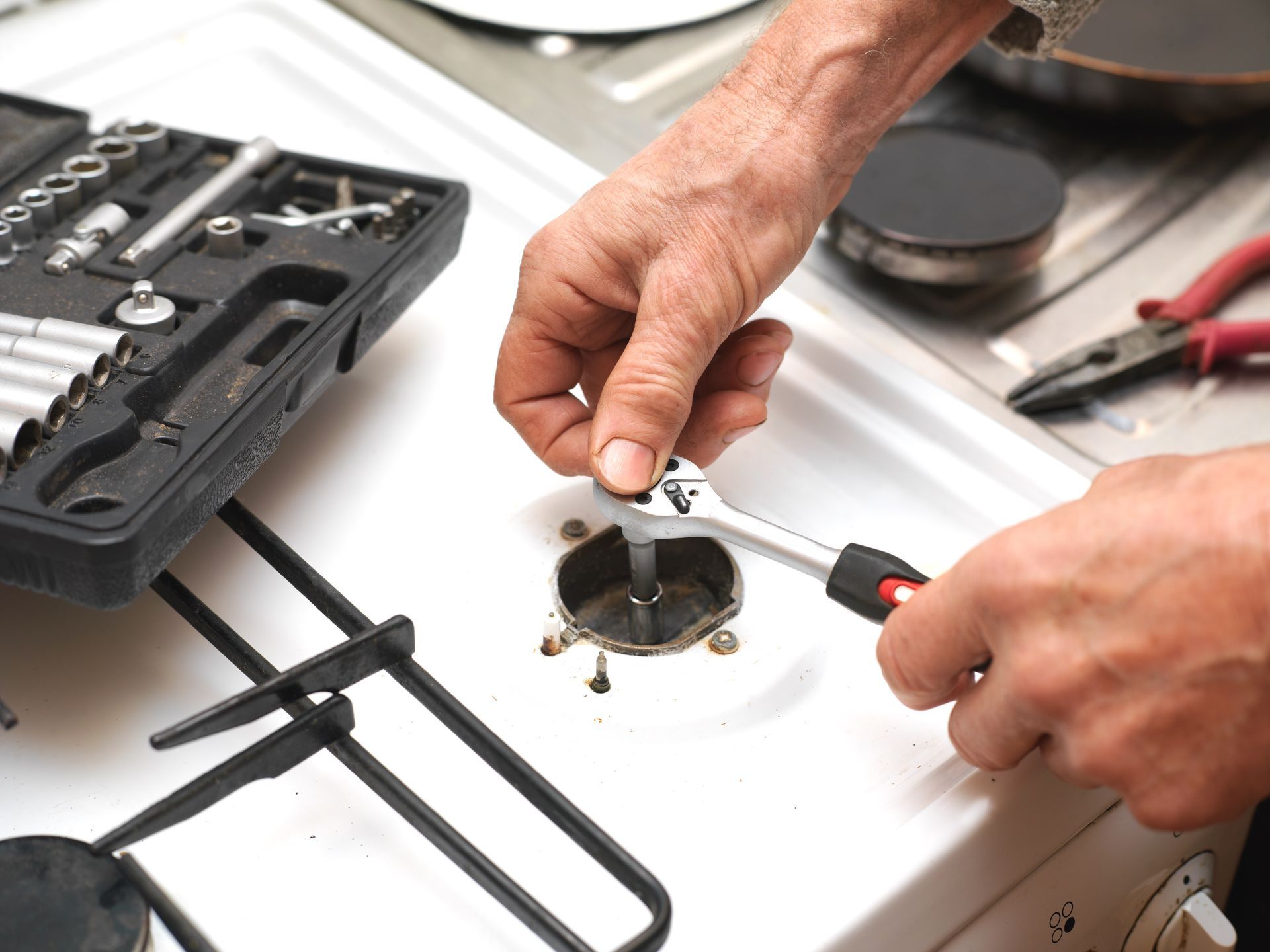 Hands using a wrench to repair a white appliance with tools on a workbench