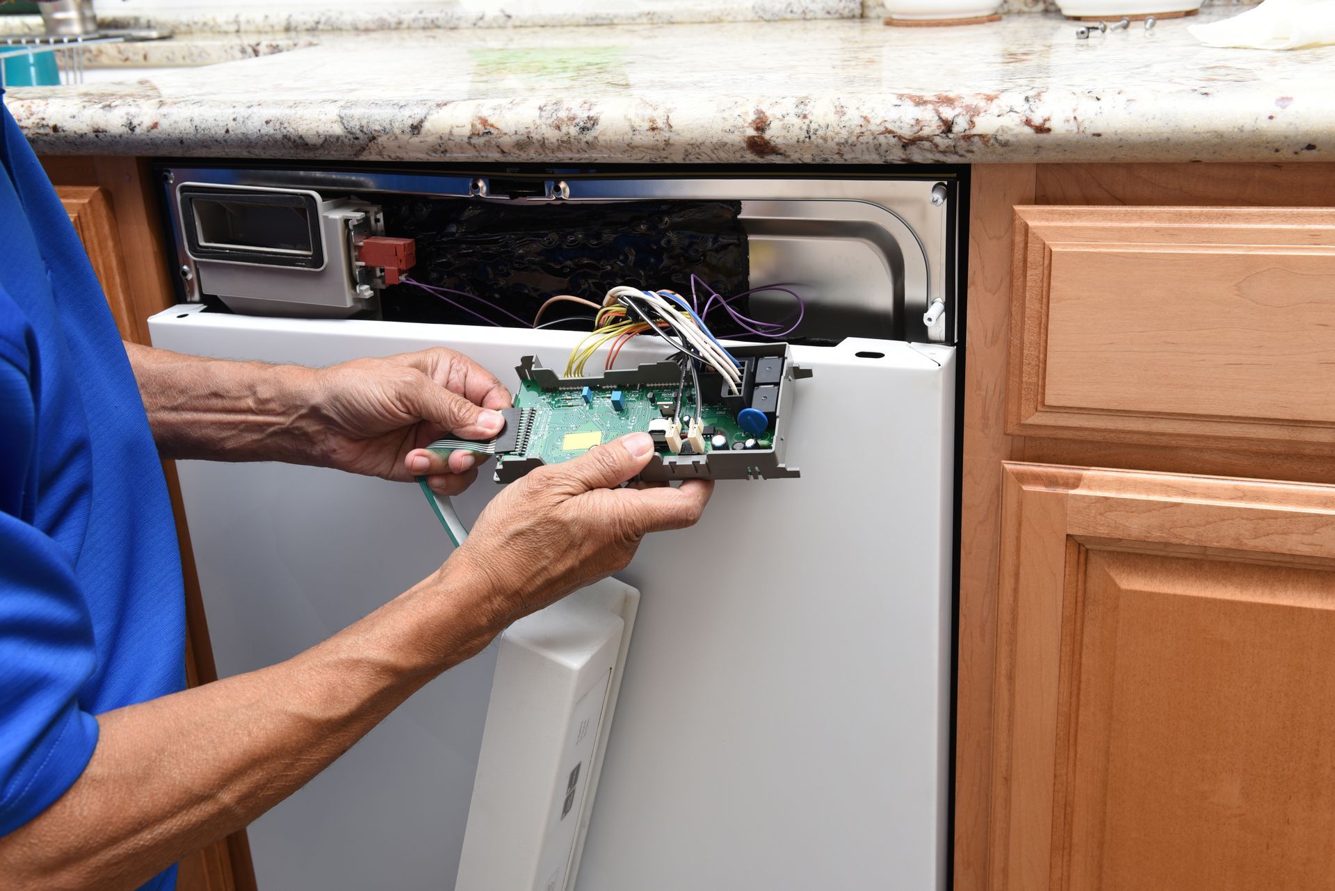 Technician repairing a dishwasher under a kitchen countertop with tools and exposed wiring