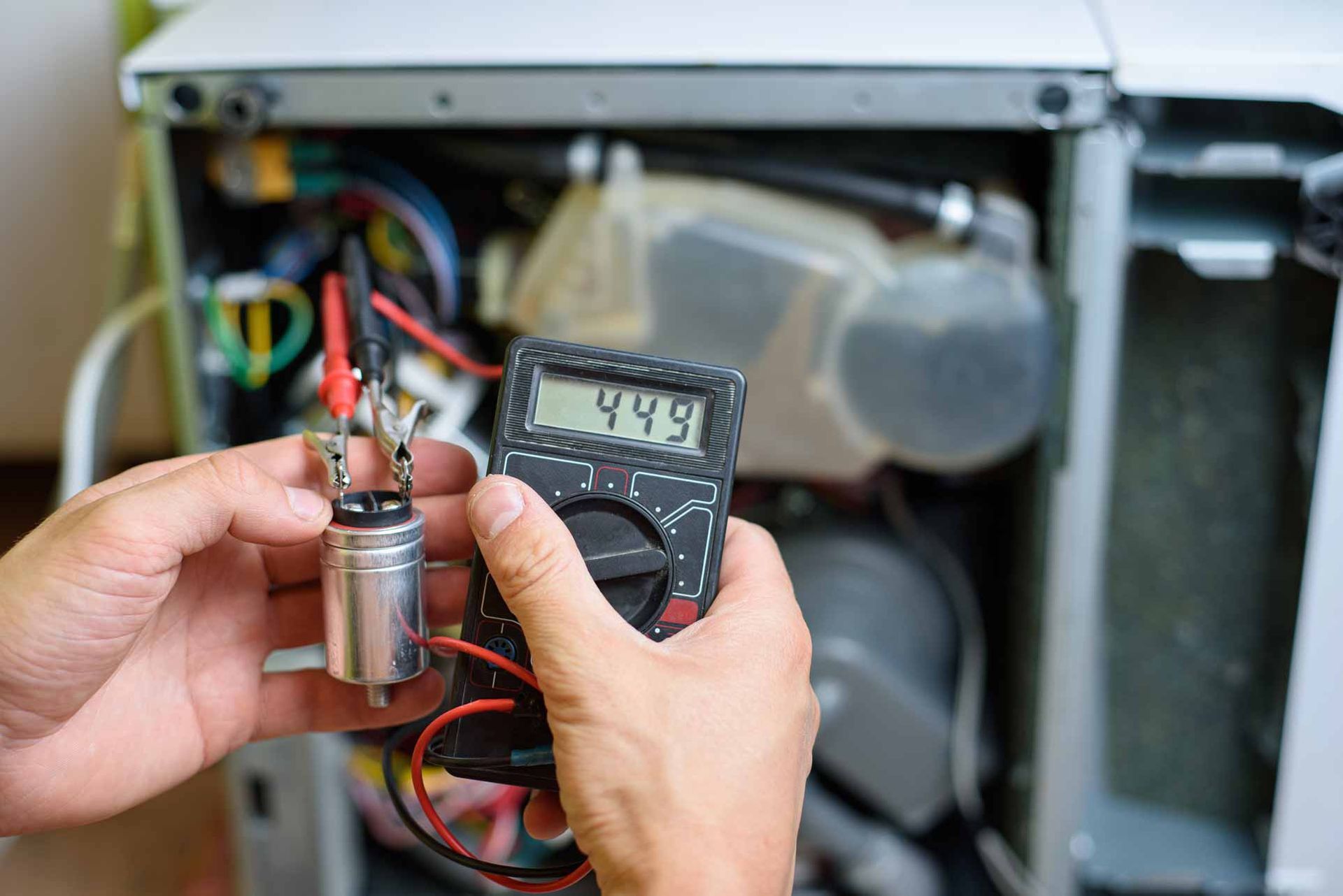 Hands holding a digital meter and a small cylindrical component inside an open machine cabinet