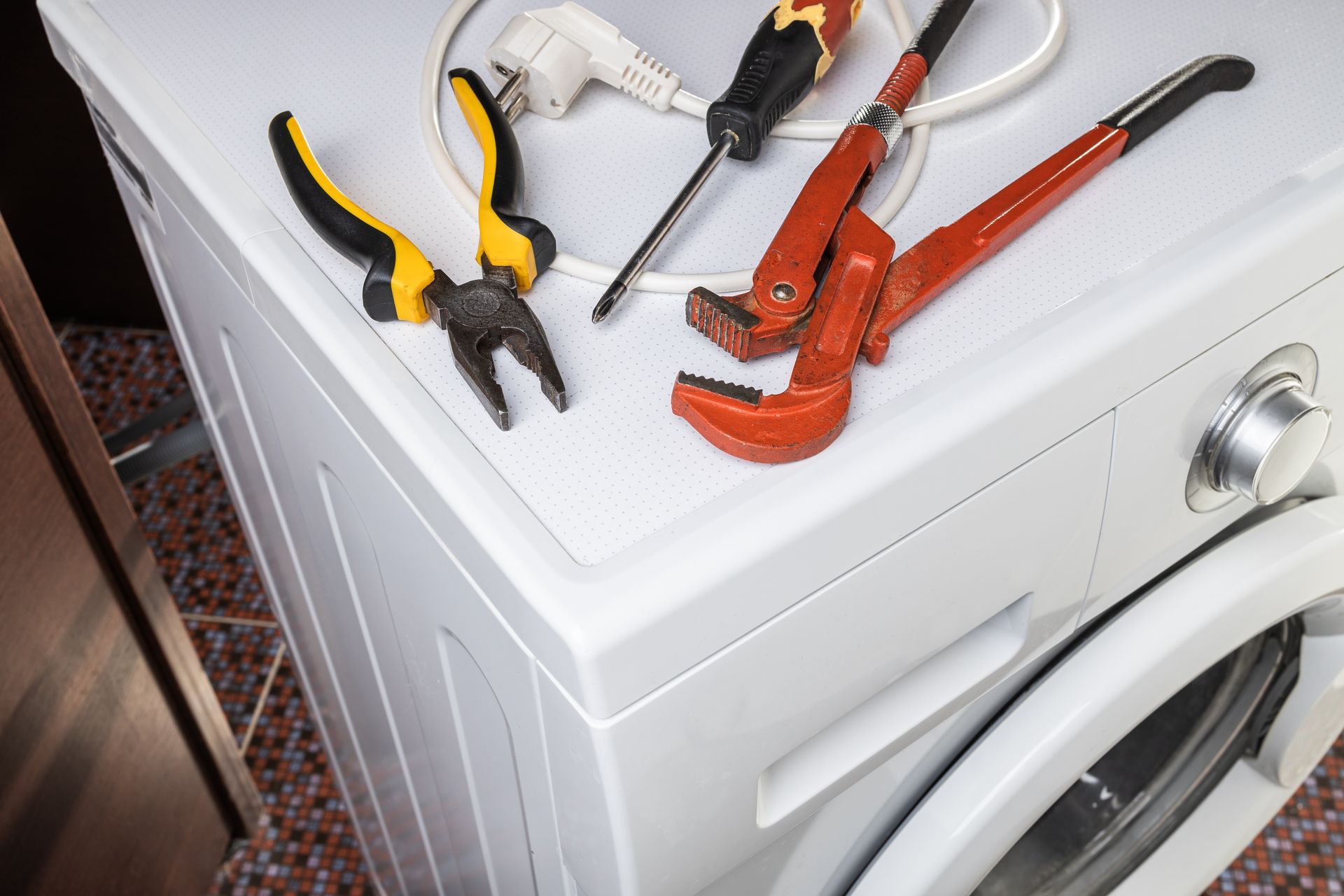 Tools on top of a white washing machine: pliers, screwdriver, wrench, and orange pipe wrench.