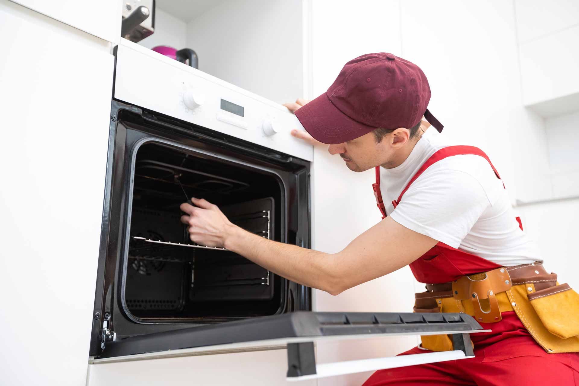 Technician in red apron and cap cleaning the inside of a built-in oven in a white kitchen