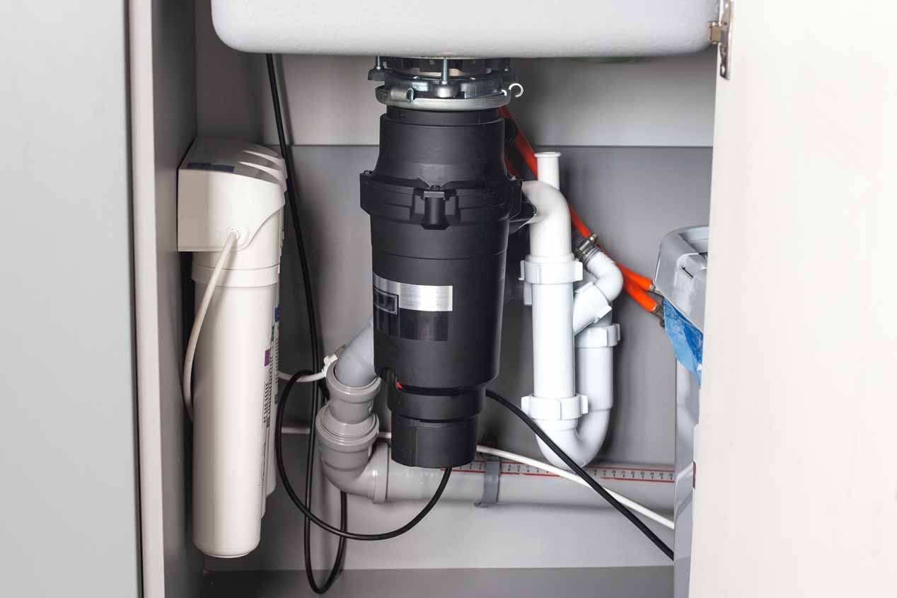 Under-sink plumbing with black garbage disposal, white pipes, and exposed water lines