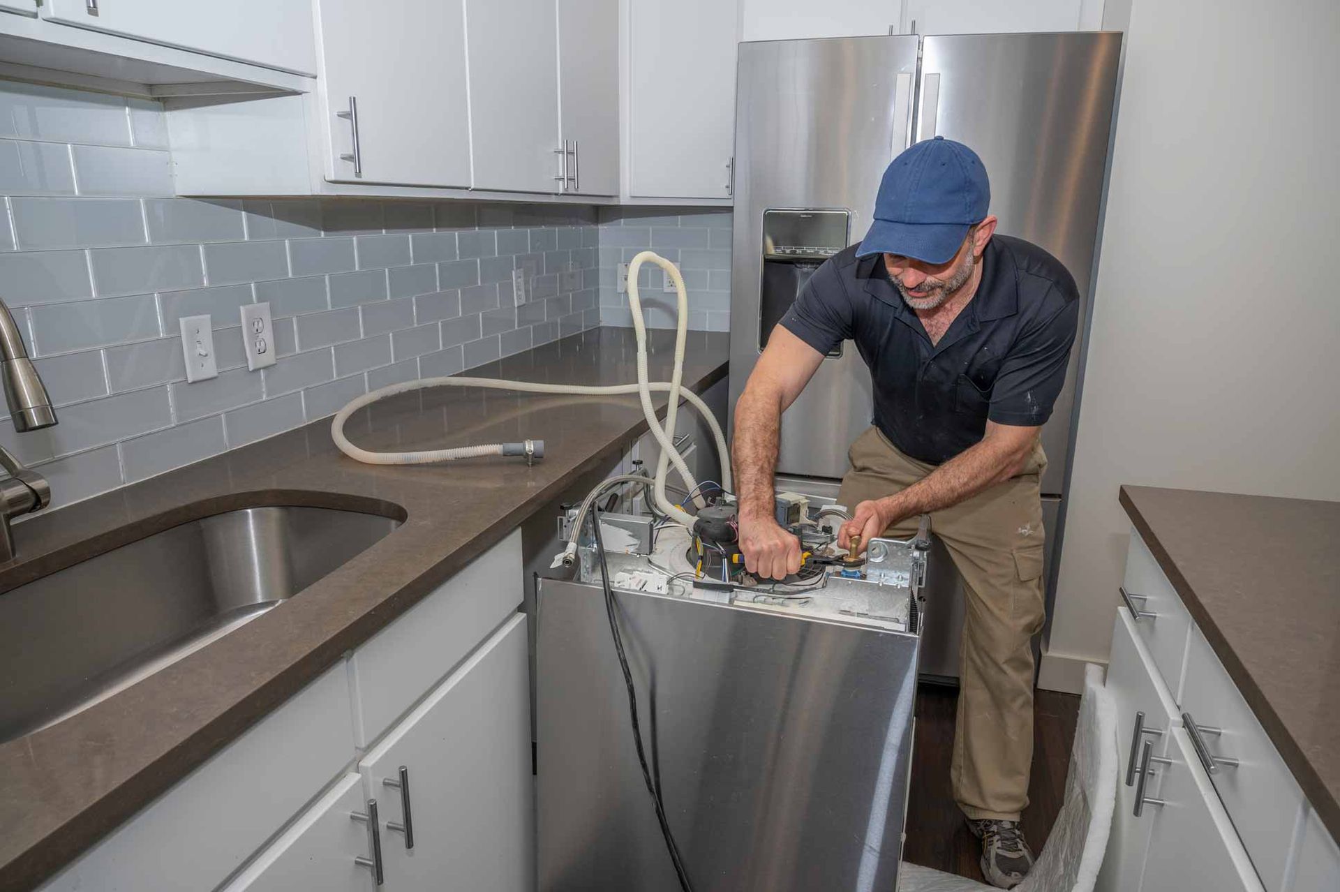 Technician repairing a dishwasher in a modern kitchen, with cabinets, sink, and stainless-steel appliances nearby