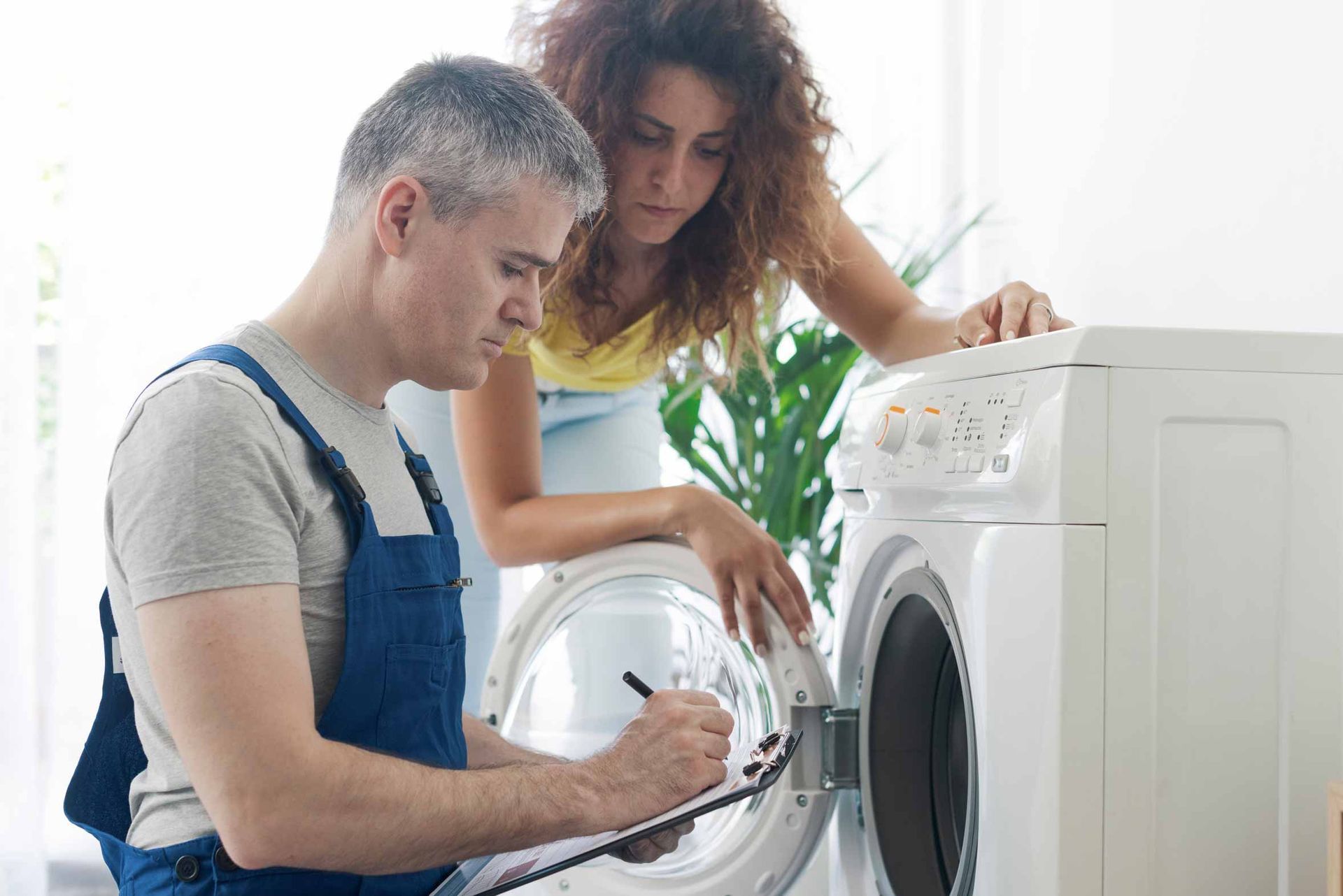 Two people repairing a front-loading washing machine in a bright laundry room