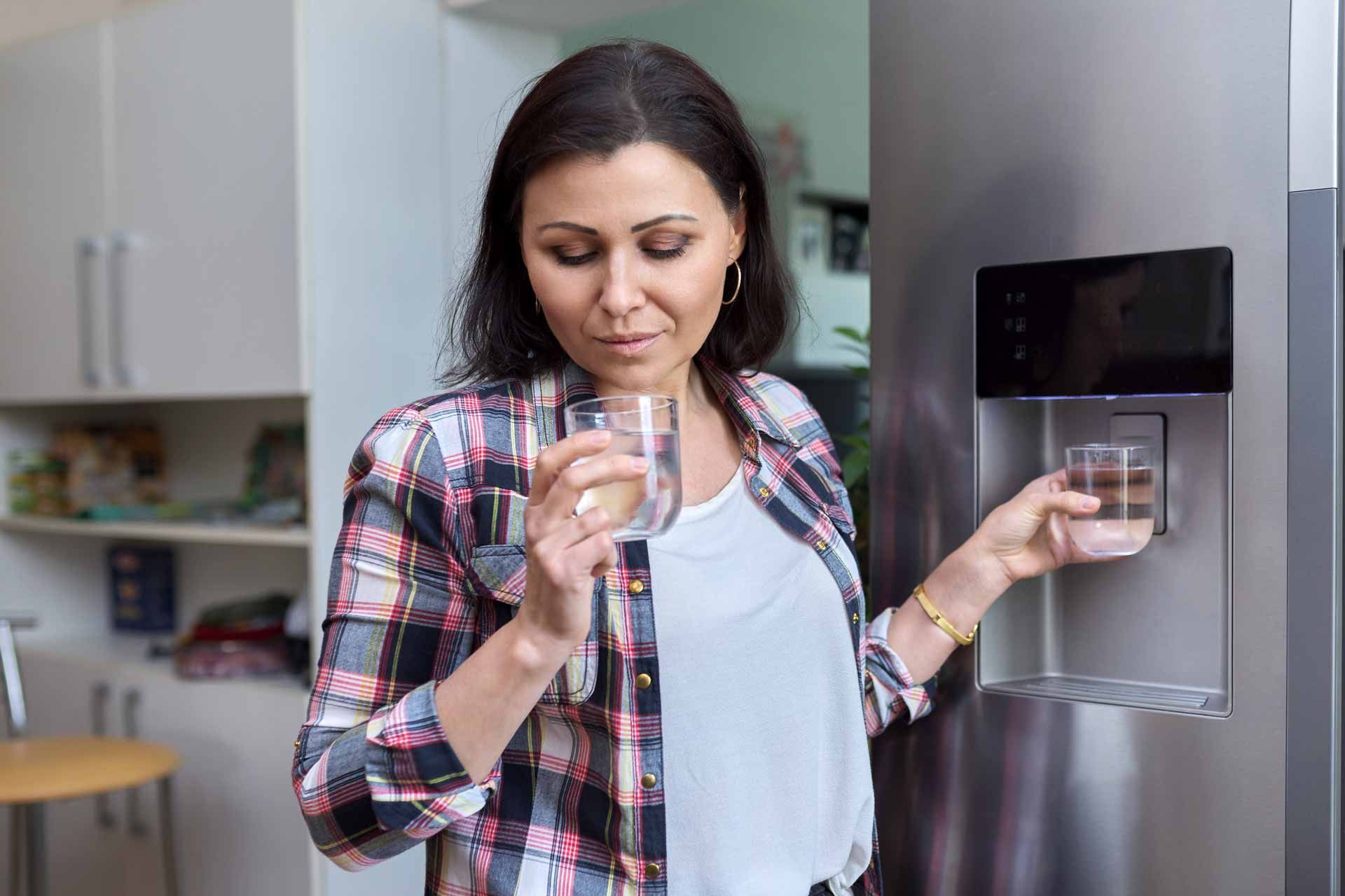 Woman drinking from a glass by a stainless steel refrigerator, holding the water dispenser tray.