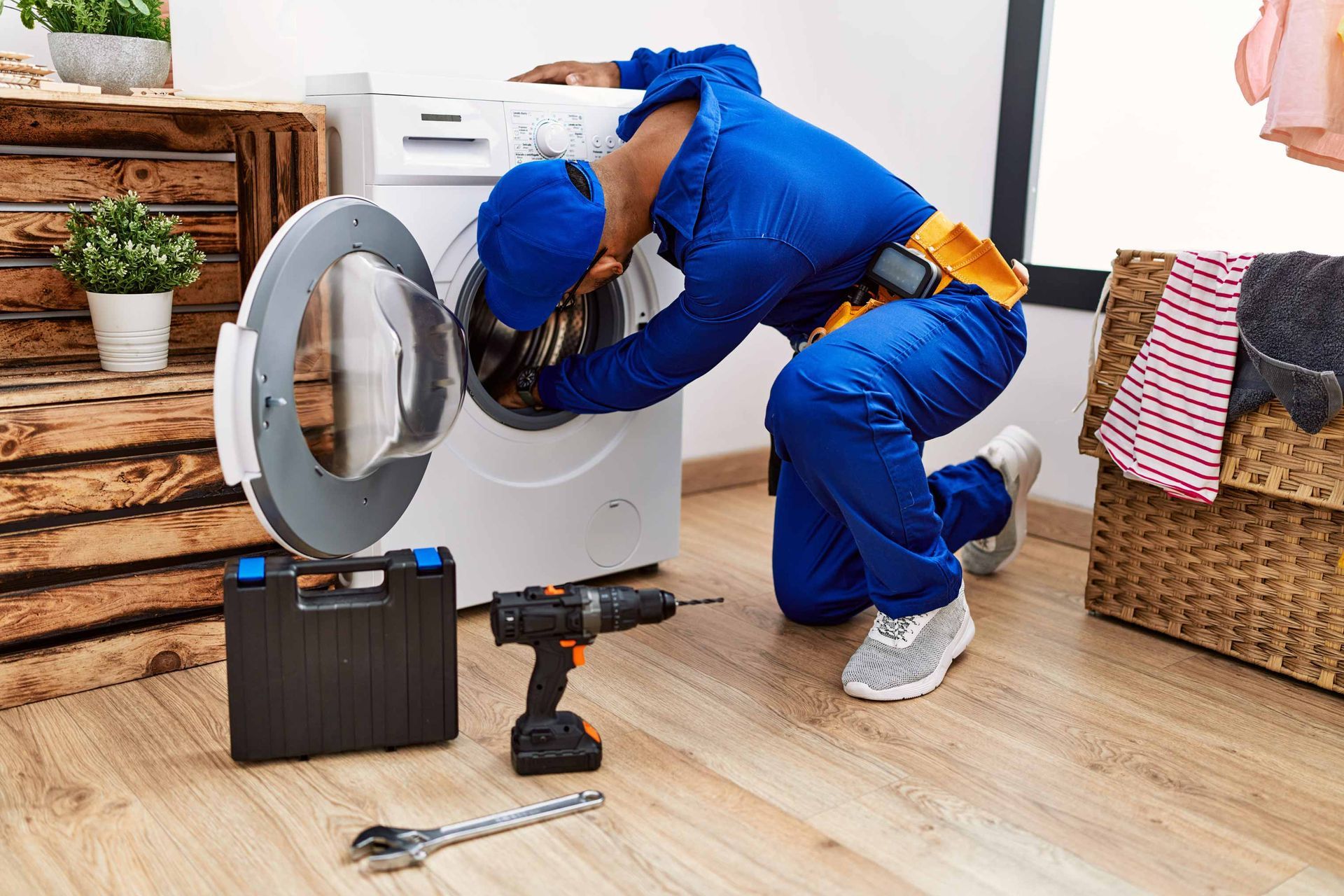 Plumber in blue coveralls repairing a washing machine in a laundry room, with tools on the floor.