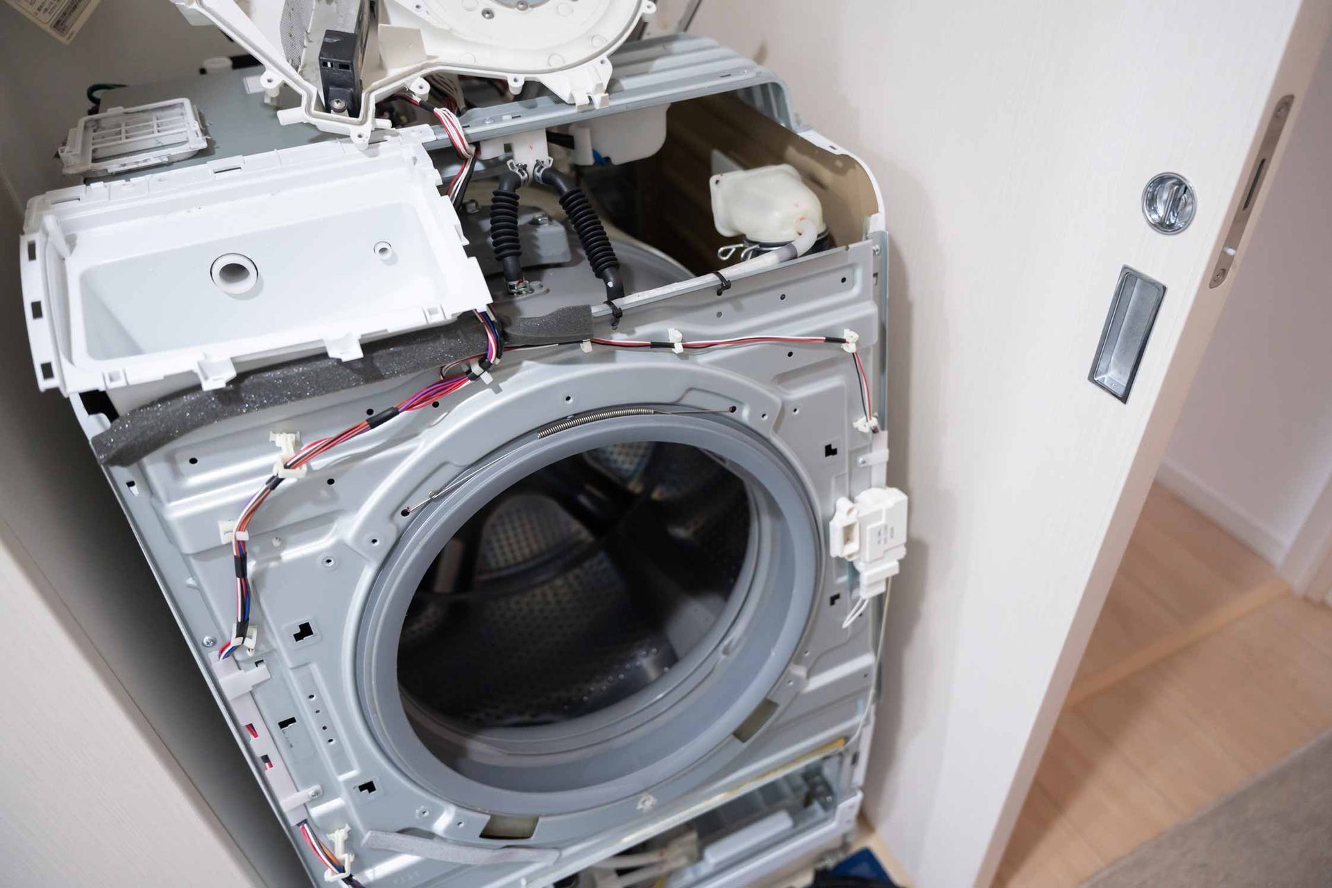Front-loading washing machine with its front panel removed, exposing internal parts in a laundry room.