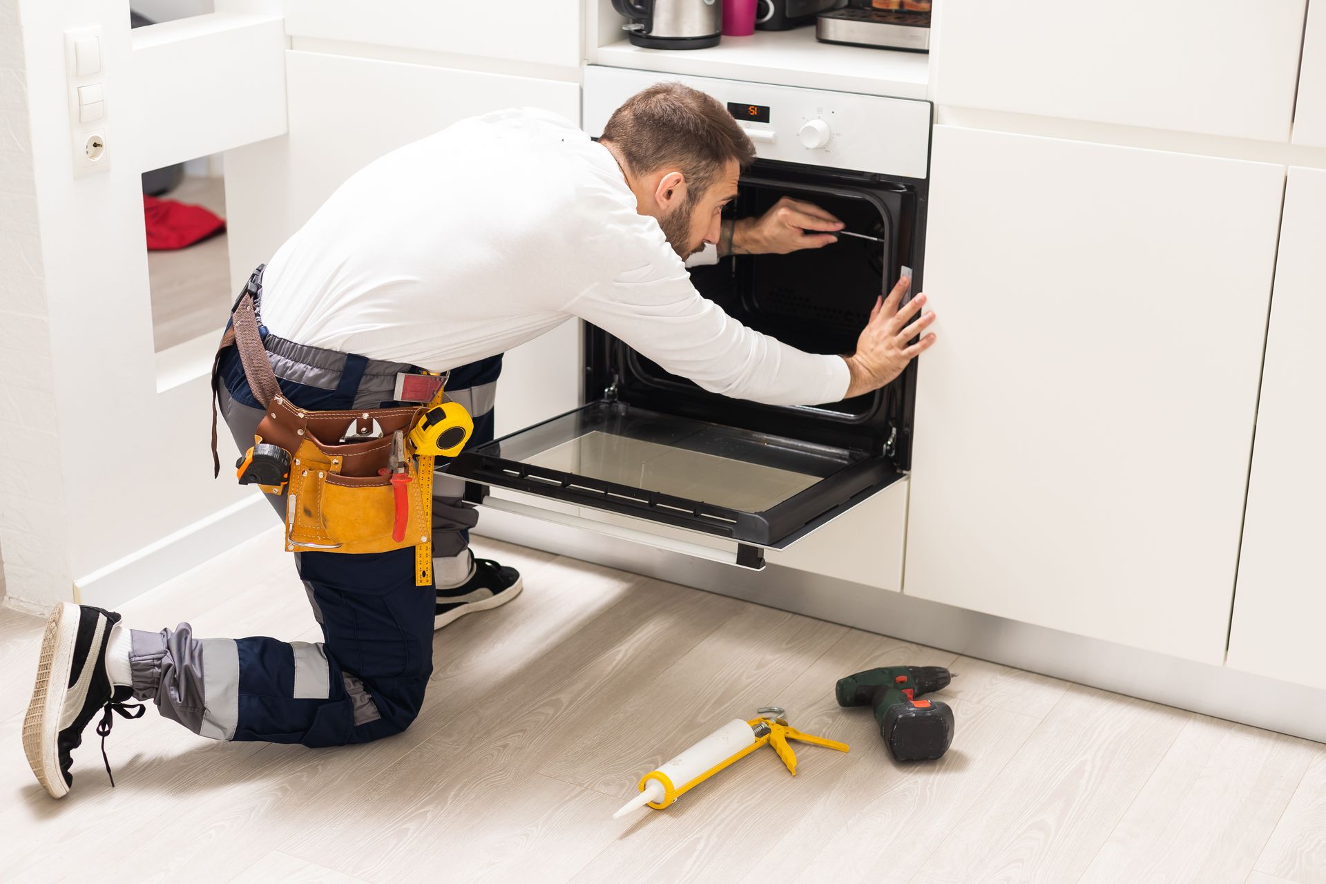 Technician kneeling to inspect a built-in oven in a bright kitchen, with tools on the floor.