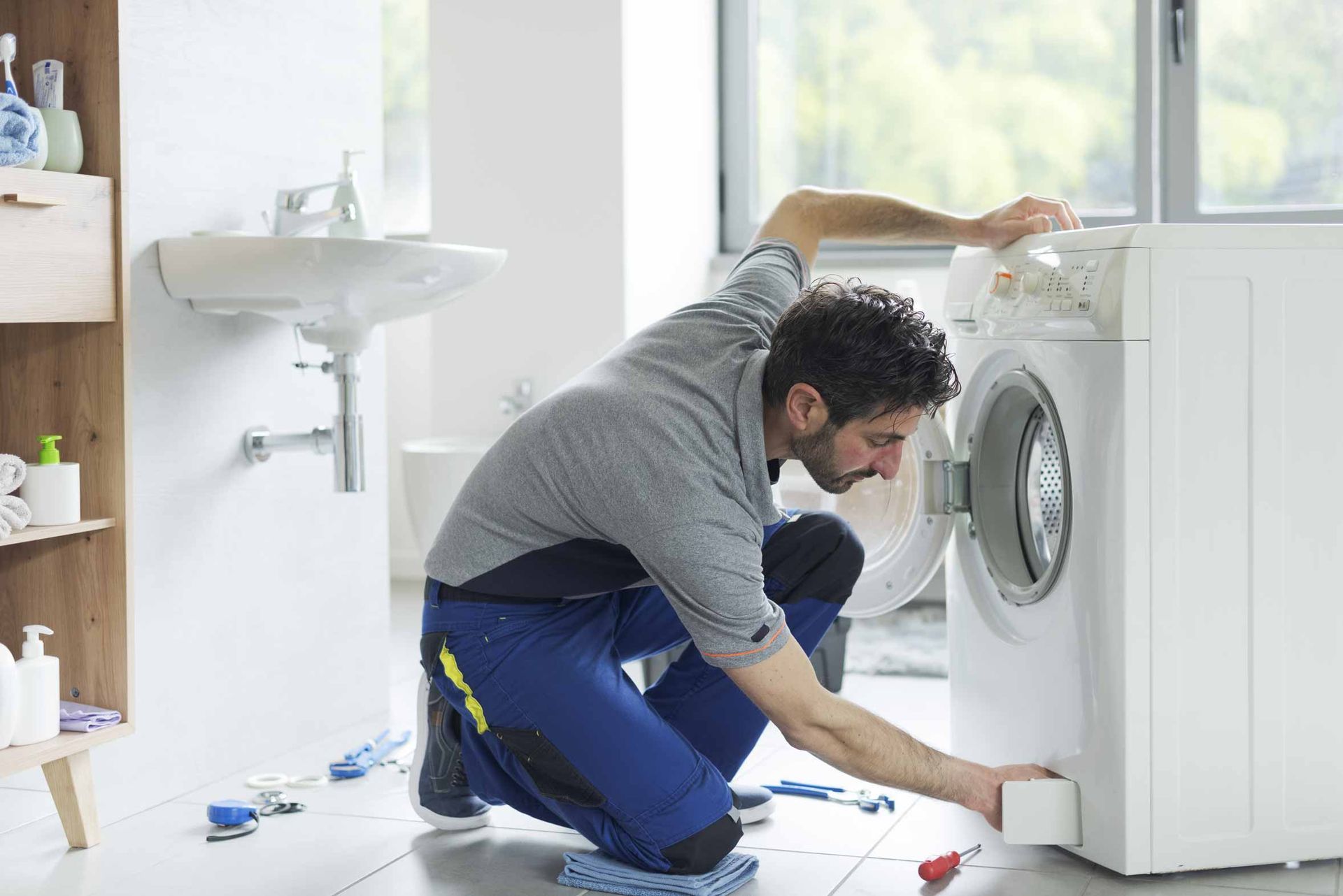Man crouching to repair a front-loading washing machine in a bright laundry room
