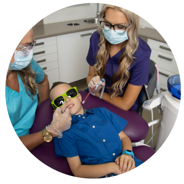 A boy wearing sunglasses is getting his teeth examined by two dentists