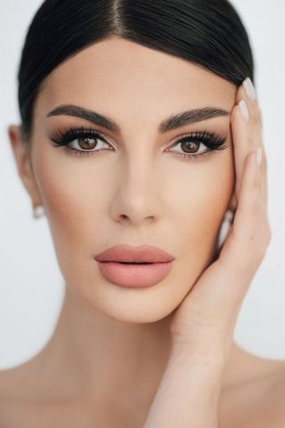 Close-up portrait of a woman with sleek dark hair and neutral makeup, resting her hand on her face.