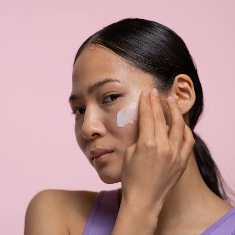 Woman applying white cream to her cheek against a pink background