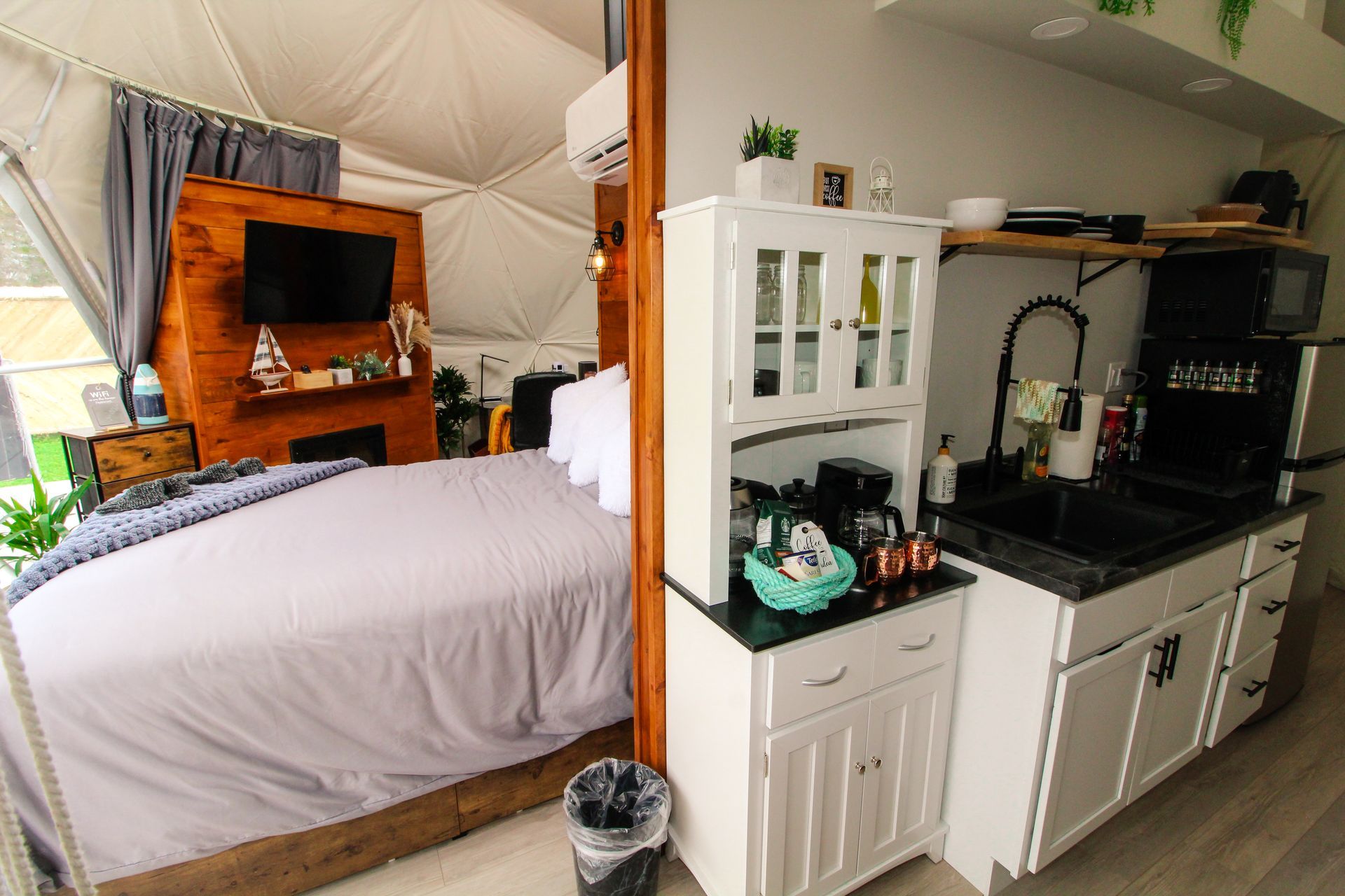 Interior of a dome-shaped tiny home with a kitchen, bed, and TV. The color palette is white, gray, and wood.