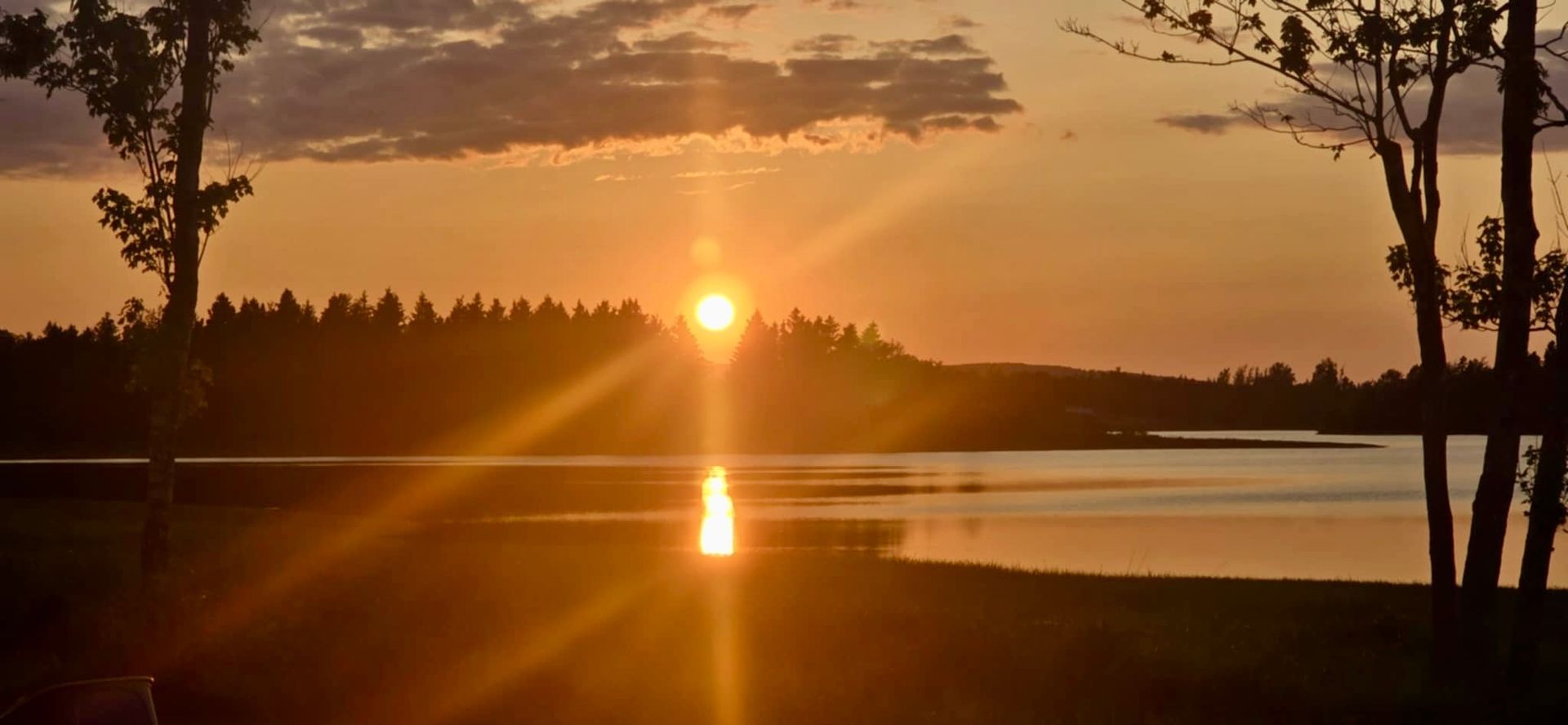 Sunset over a lake; sun's rays reflect on water, trees frame the view, sky glows orange.