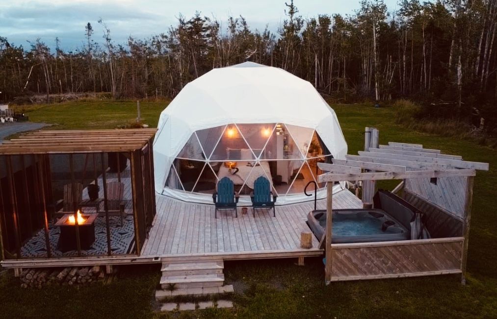 White geodesic dome with wooden deck, fire pit, and hot tub, set in a grassy area with trees.