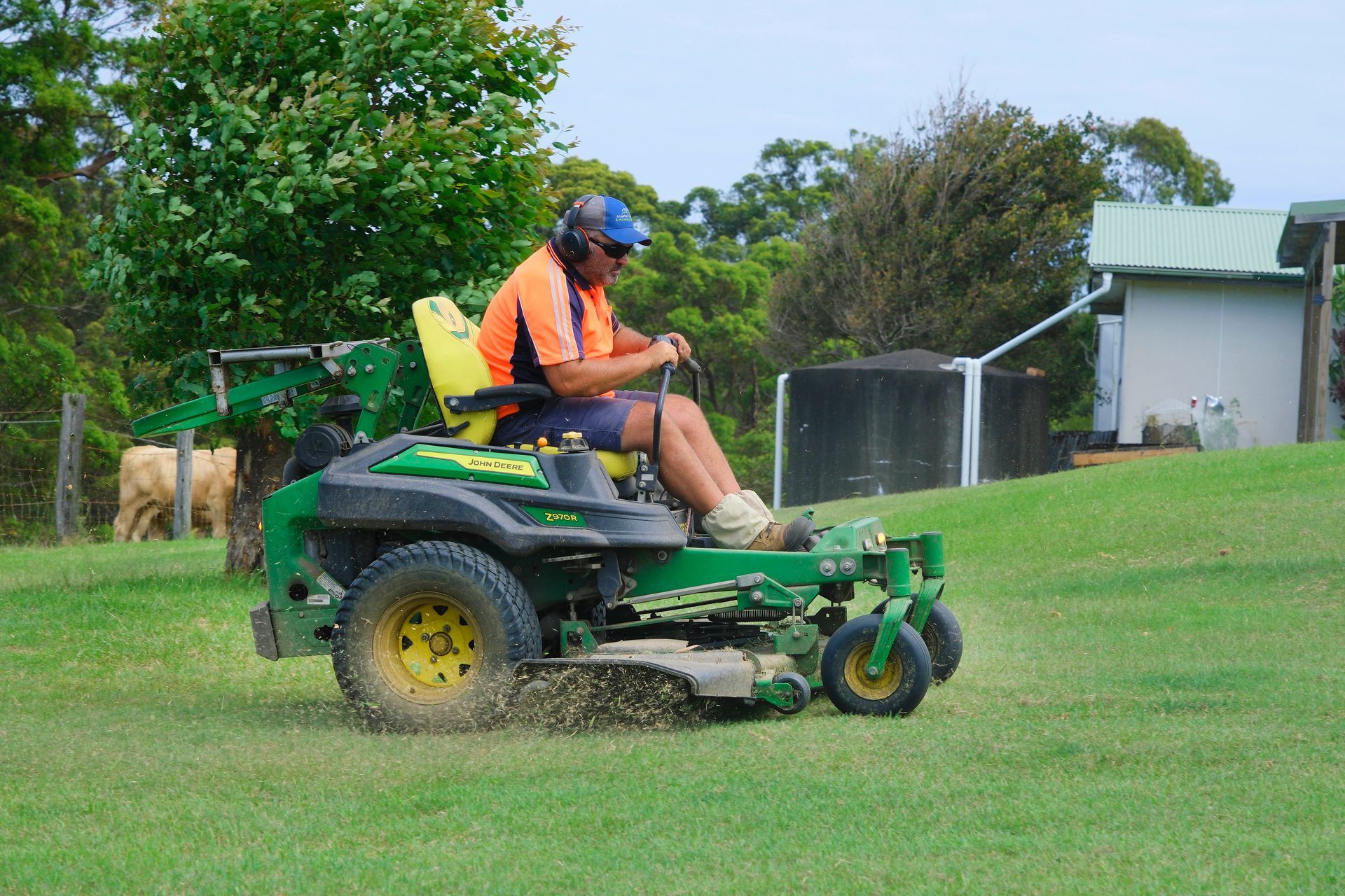 A person wearing a bright orange shirt and cap operates a green riding lawn mower on a grassy field near a white shed. Coastal Slashing & Mowing Services In Tallwoods Village, NSW