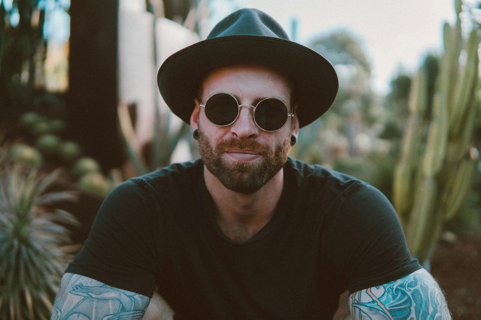 A man wearing a hat and sunglasses is sitting in front of a cactus.