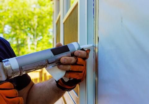 Person using a caulk gun to seal a door frame. Wearing orange work gloves. Outdoors.