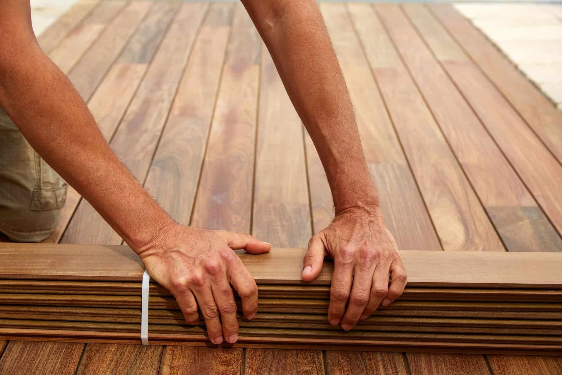 Person's hands holding a stack of wooden planks. Some planks are laid on the ground.