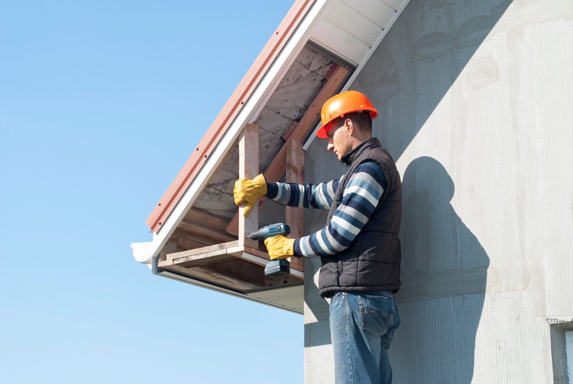 Construction worker in orange hard hat drills wood on building's overhang.