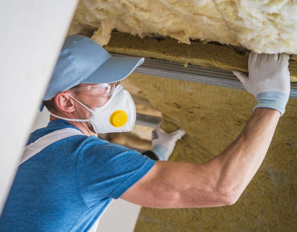 Man installing insulation in a ceiling, wearing a mask, safety glasses, and gloves.