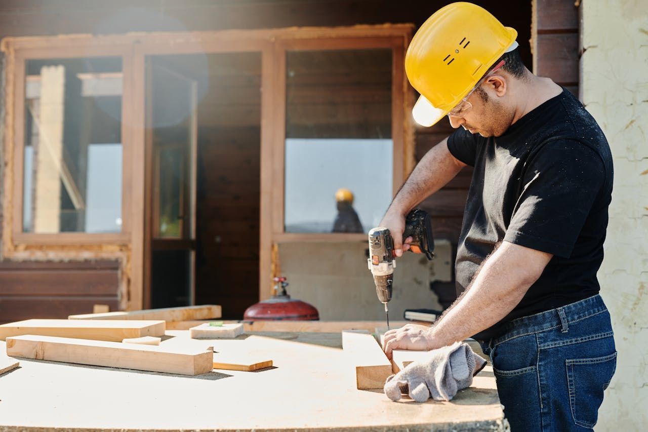 Construction worker in a yellow hard hat drills wood on a table outdoors near a building with windows.