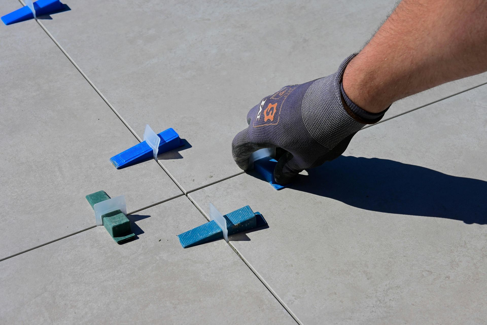 Hand with work glove using a tool to adjust tile leveling clips on a tiled surface.