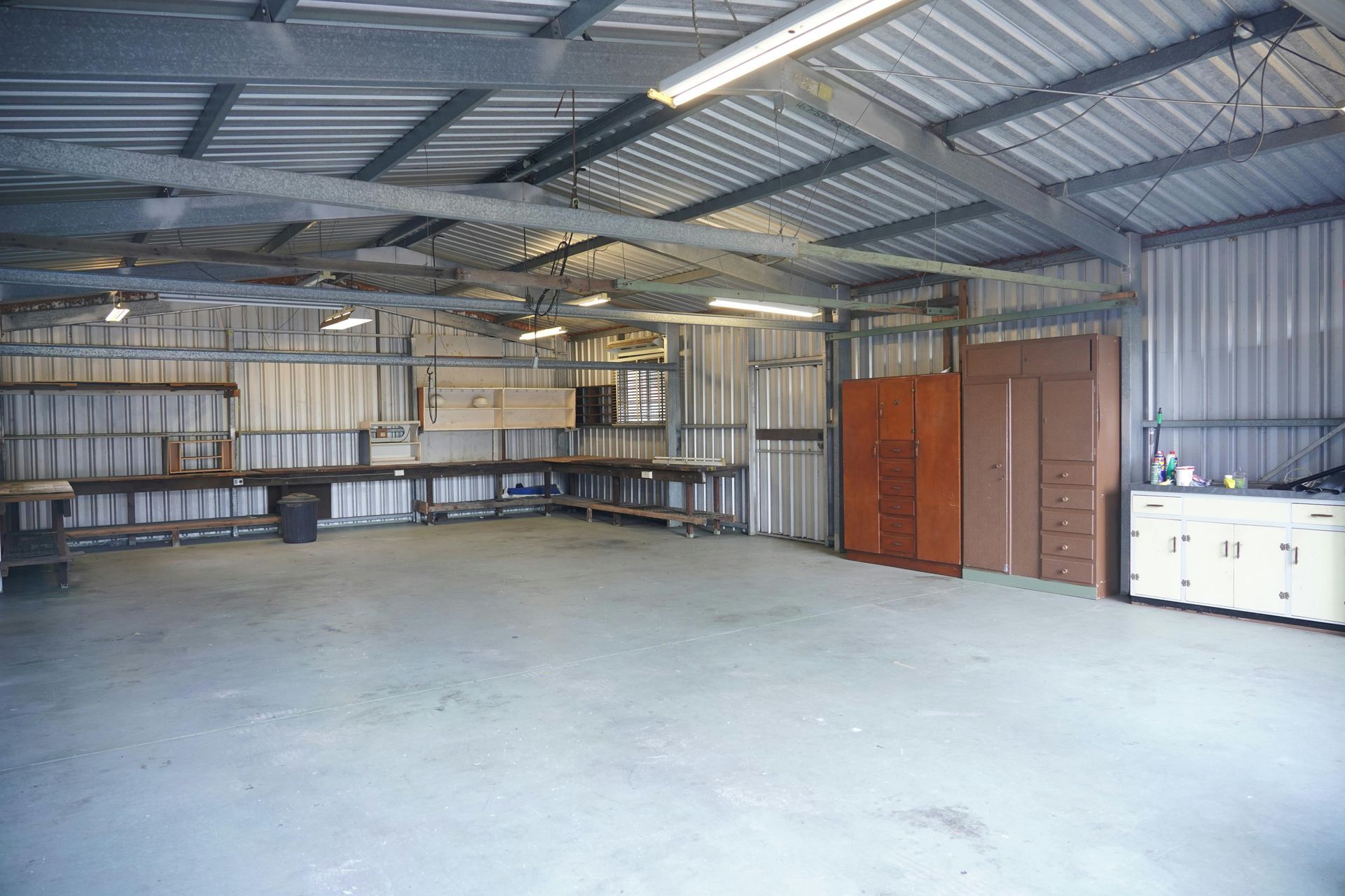 Interior of a metal workshop with a concrete floor, workbenches, cabinets, and tall wooden storage units.