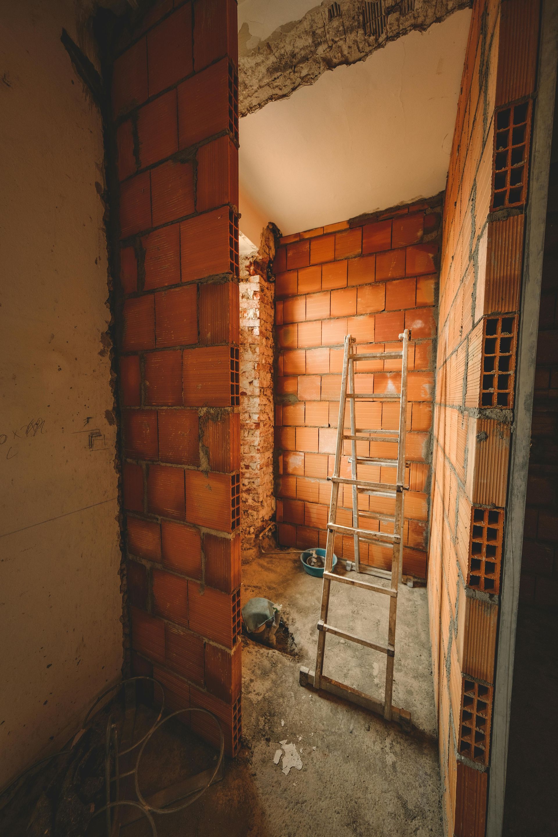 Interior of a room under construction with exposed brick walls, a ladder, and concrete flooring.