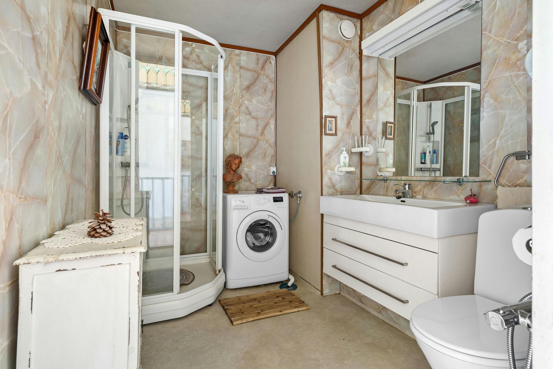 Bathroom with a white toilet, sink, and washing machine. Shower stall and marble-look walls.