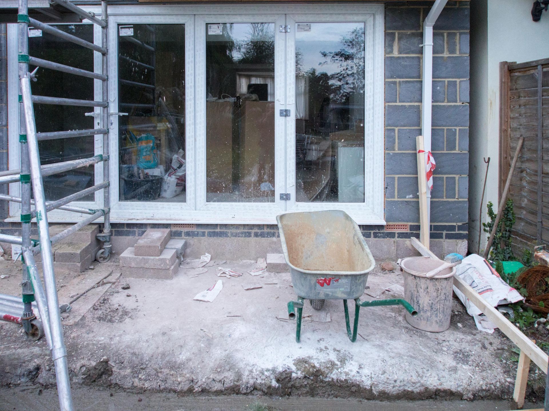 Construction site: white door, gray wheelbarrow, brick wall, ladder, debris on concrete base.