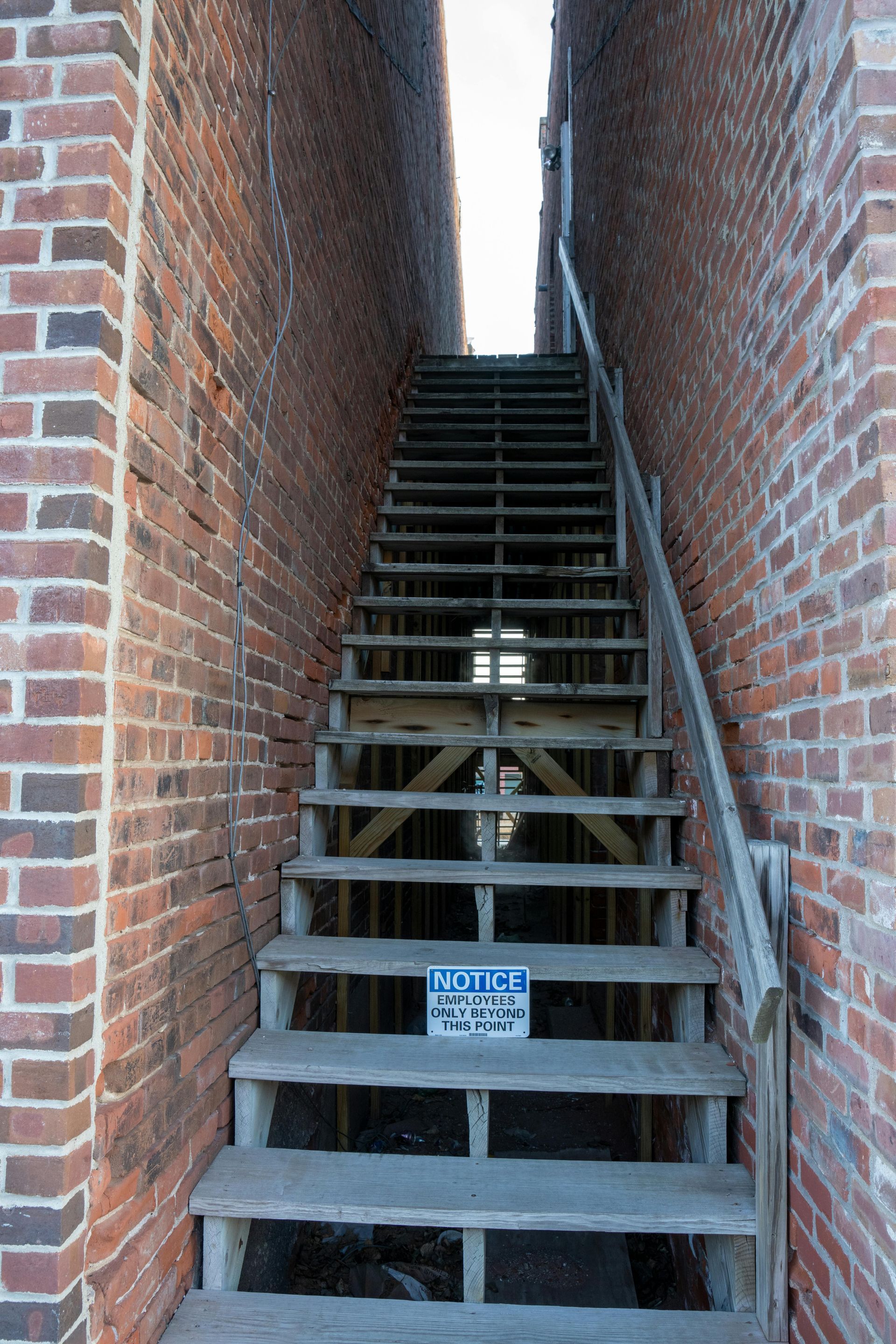 Wooden staircase between brick walls, leading upwards. Warning sign on the stairs.