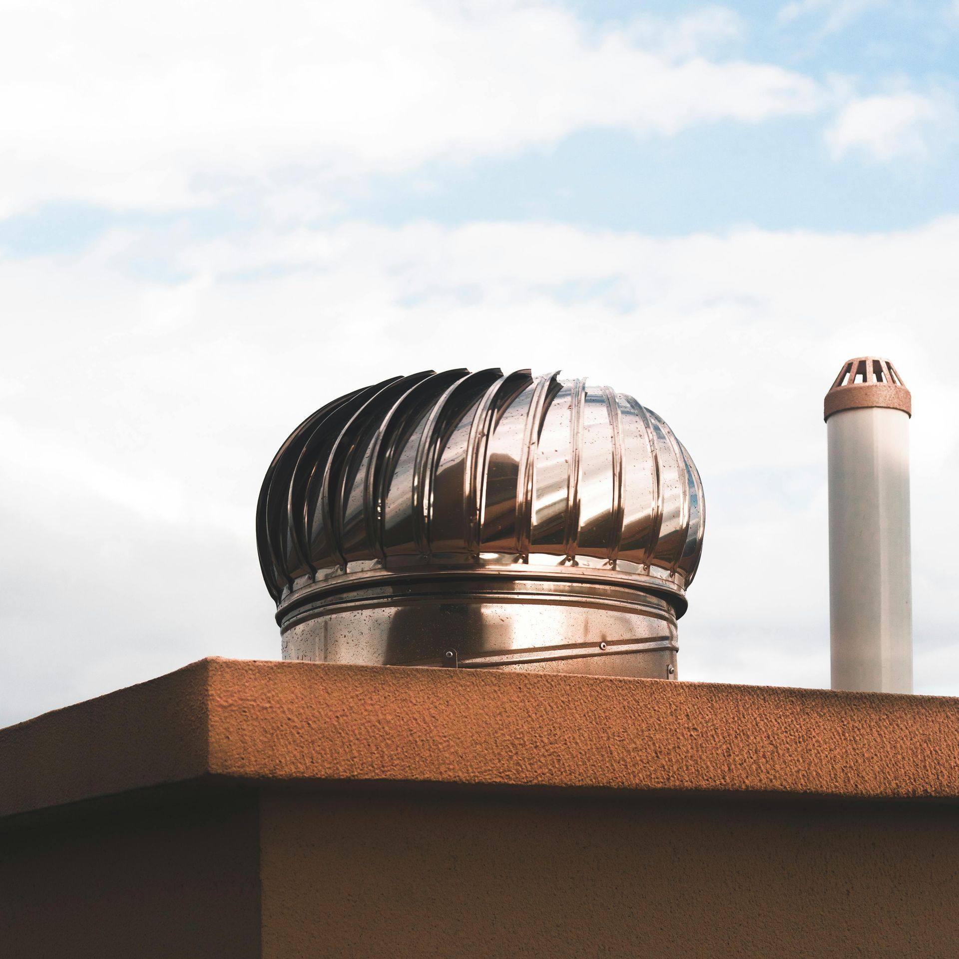 Shiny, rotating roof vent on a flat-roofed building, beside a chimney, against a cloudy sky.