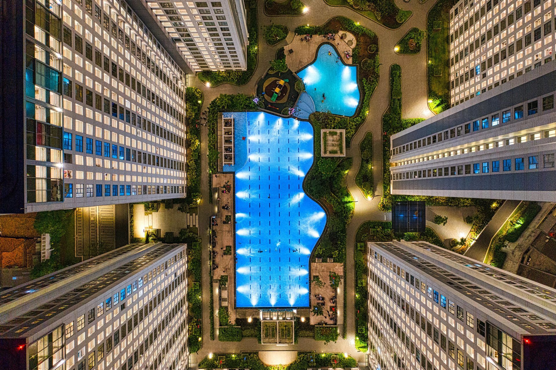 Aerial view of apartment buildings surrounding a large, illuminated swimming pool at night.