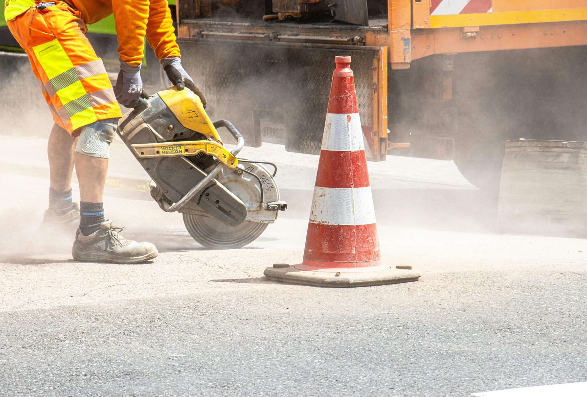 Road worker cuts asphalt with saw; cone nearby.